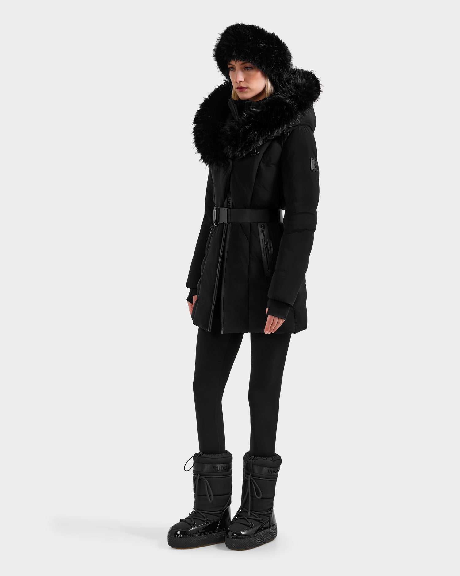 A person stands against a plain light background wearing the Rudsak MODA FF - 8125969 Black/Black belted winter coat with a large faux fur hood, black leggings, a fur hat, and black winter boots.