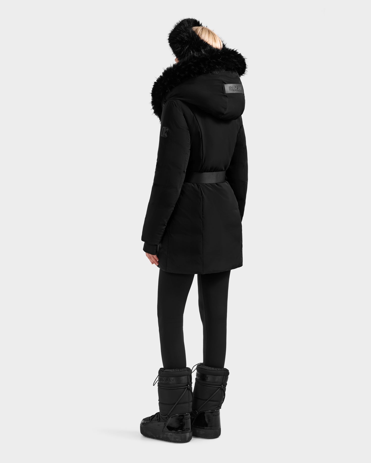 Wearing the Rudsak MODA FF - 8125969 Black/Black fitted down puffer coat with faux fur hood, black leggings, tall black boots, and a black fur headband, a person stands facing away on a plain light background.