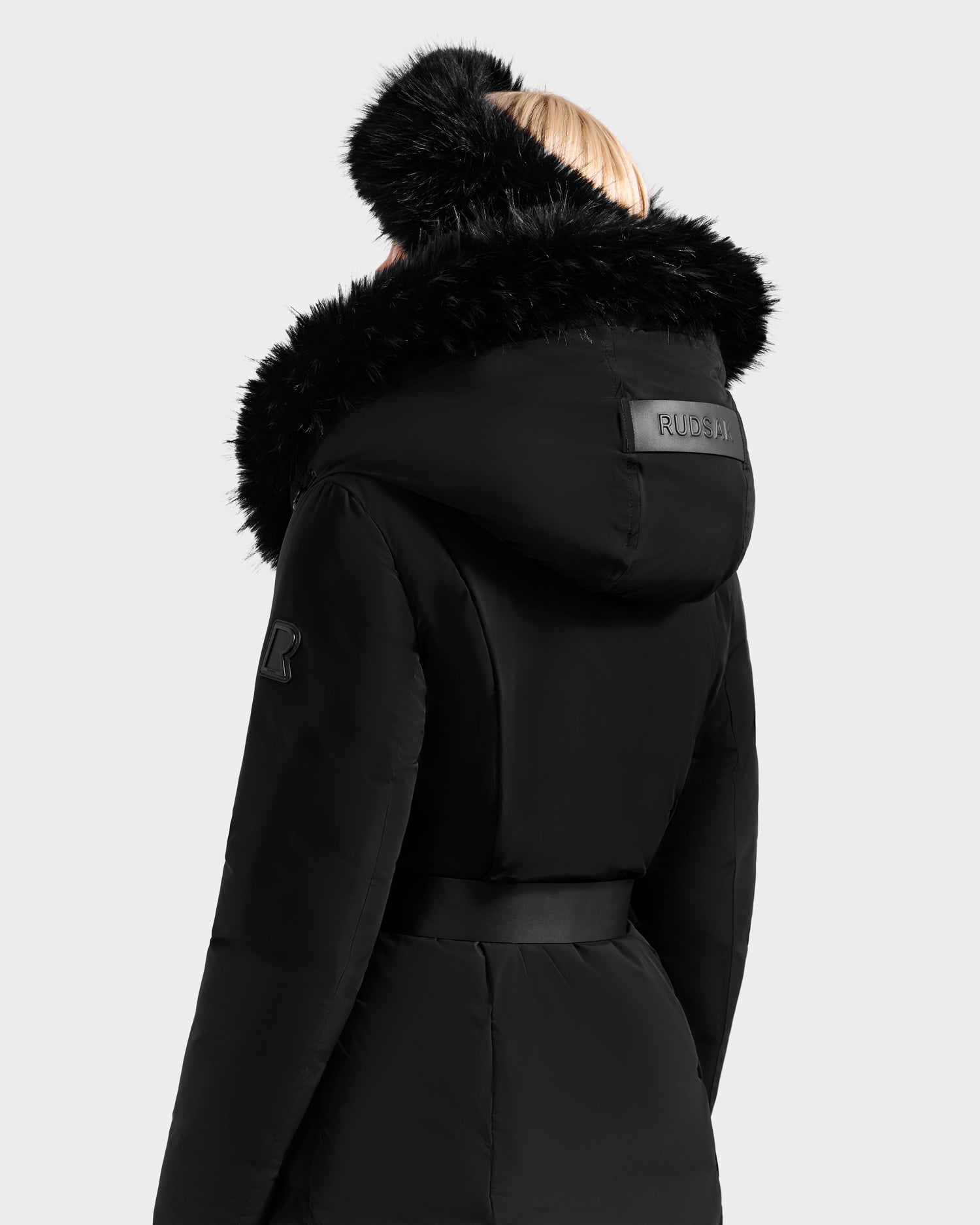 A person with blonde hair wears the Rudsak MODA FF - 8125969 Black/Black women’s fitted down puffer with a faux fur hood, black belt, and black furry earmuffs. The coat features a RUBAI label at the back against a plain light gray background.