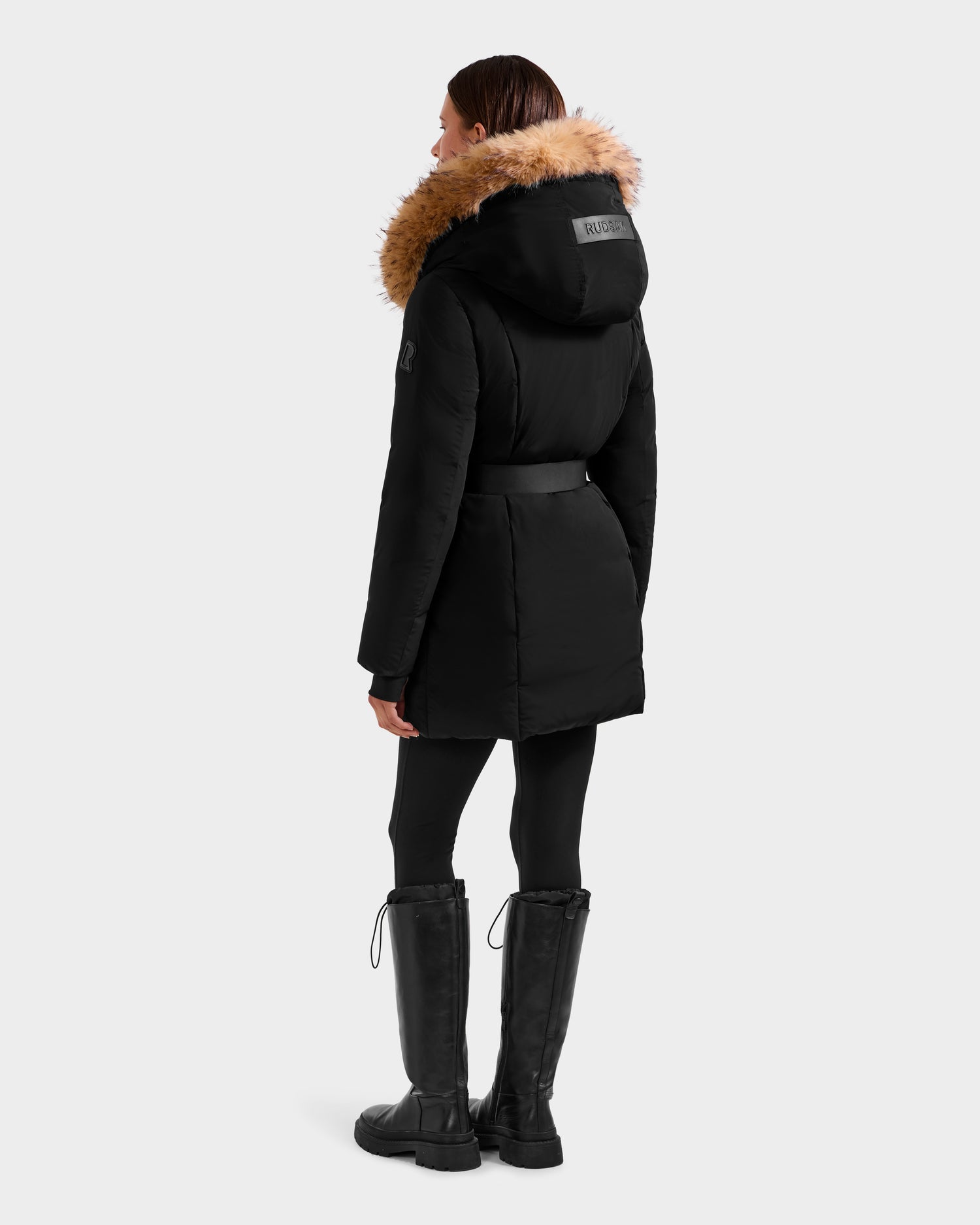 A person stands facing away, wearing the Rudsak MODA FF - 8125969 Black/Natural women’s fitted down puffer with a removable belt and brown faux fur hood, paired with black leggings and knee-high black boots against a light gray background.