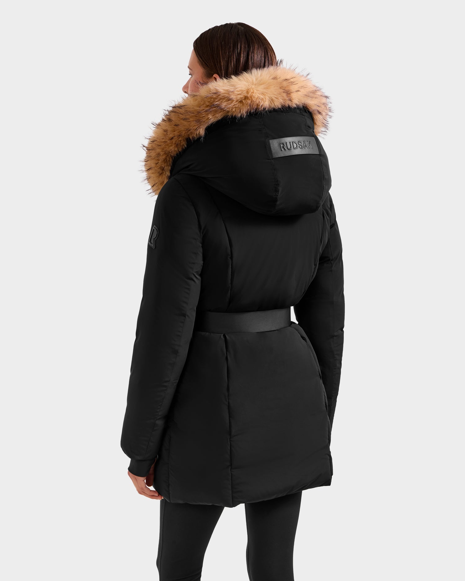 A person with straight brown hair is shown from the back wearing the Rudsak MODA FF - 8125969 Black/Natural women’s fitted down puffer, featuring a faux fur hood, removable belt, detailed stitching, and a visible RUDSAK logo patch on the hood.