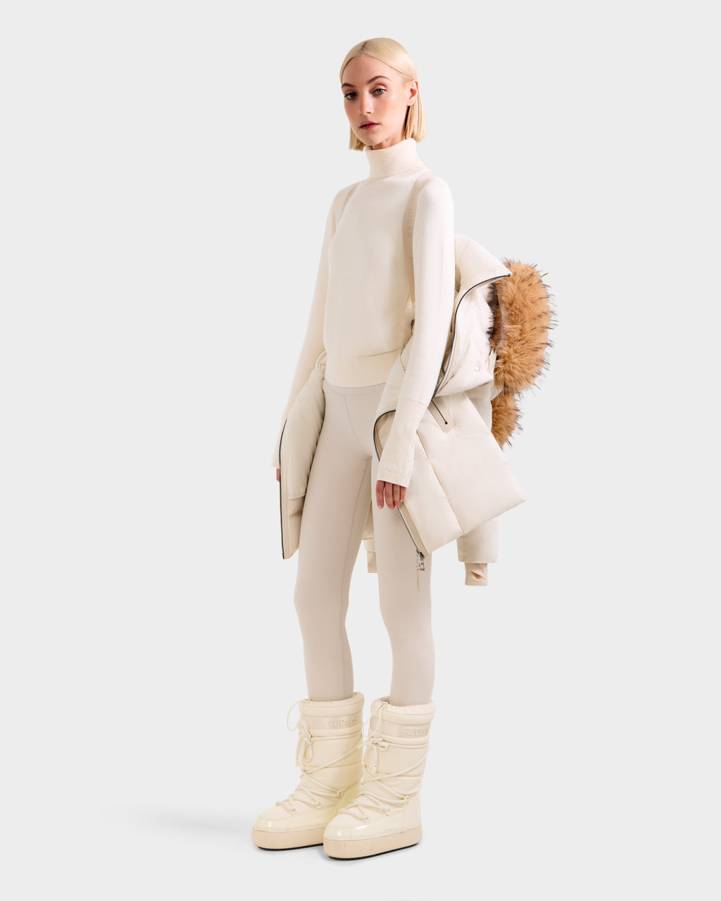 A woman in a cream turtleneck sweater, fitted pants, and puffer boots holds the Rudsak MODA FF - 8125969 Vanilla women’s fitted down puffer with faux fur hood over her shoulder, set against a light gray background.