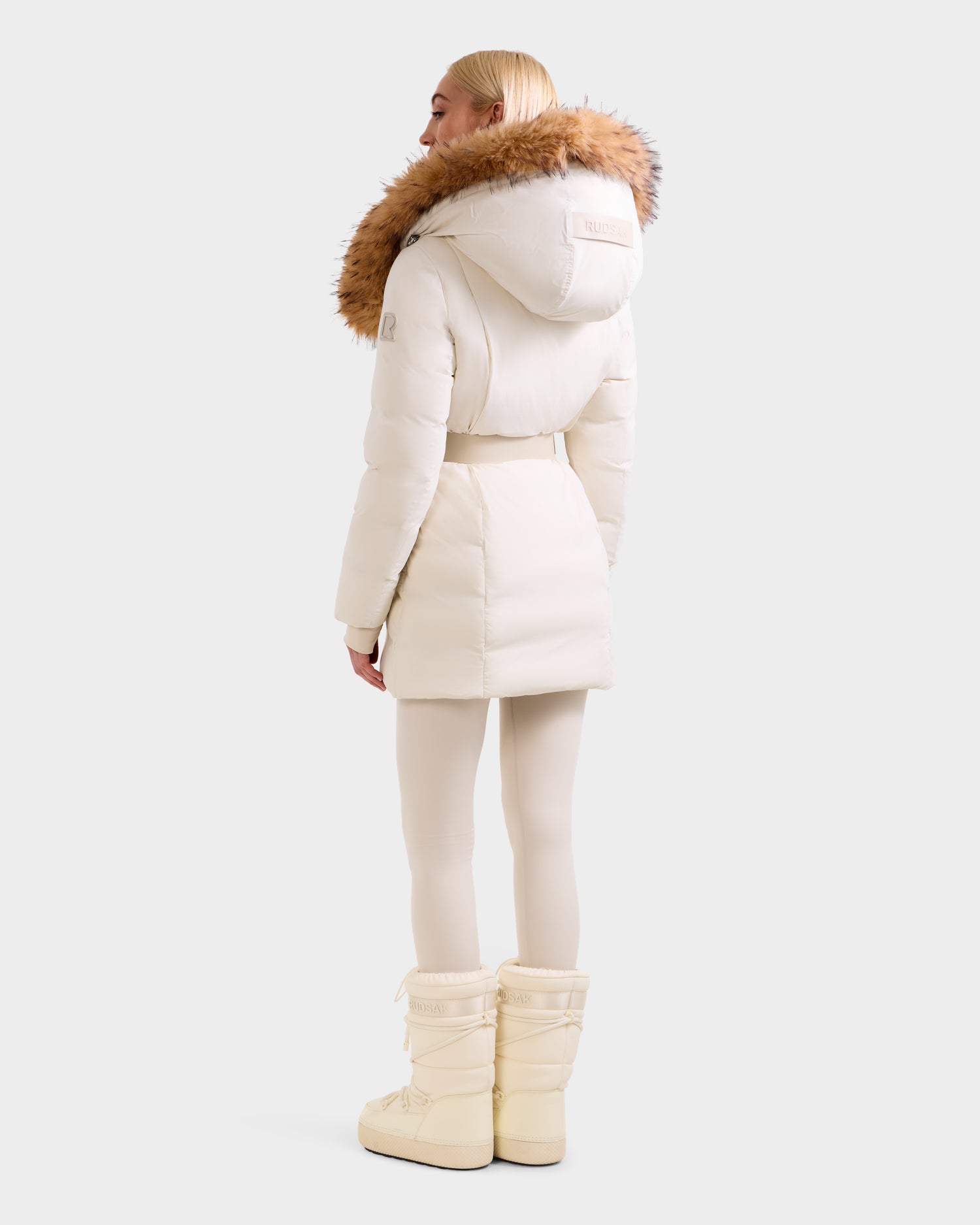 A person with straight blonde hair is seen from behind, wearing the Rudsak MODA FF - 8125969 Vanilla women’s fitted down puffer coat with faux fur hood, paired with white leggings and snow boots against a plain background.