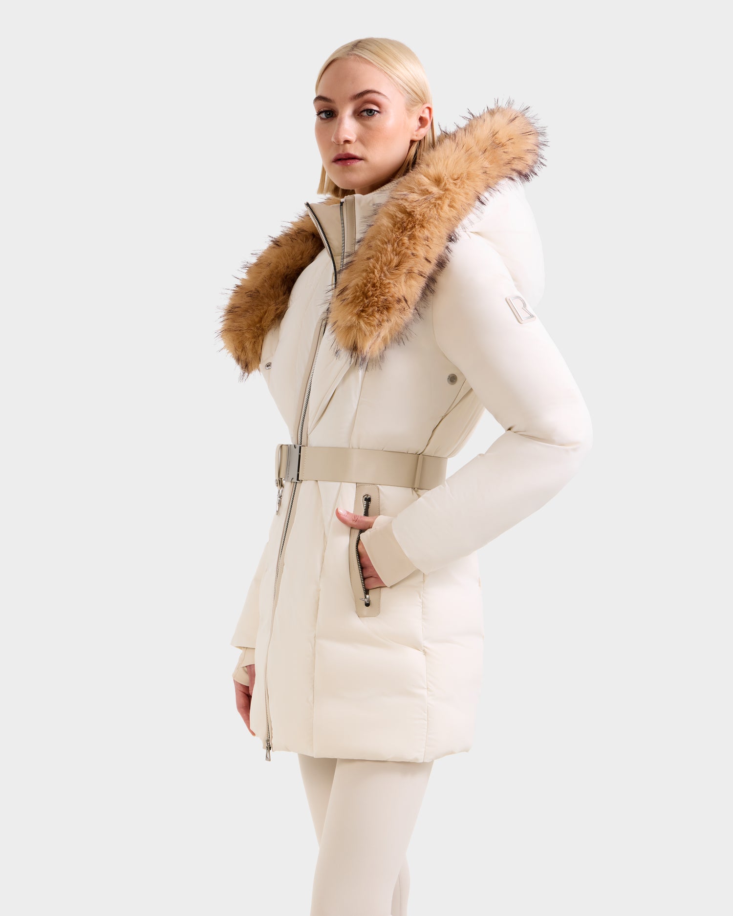 A woman wearing the Rudsak MODA FF - 8125969 Vanilla women's fitted down puffer coat with a belted waist and large faux fur hood stands against a plain light gray background.