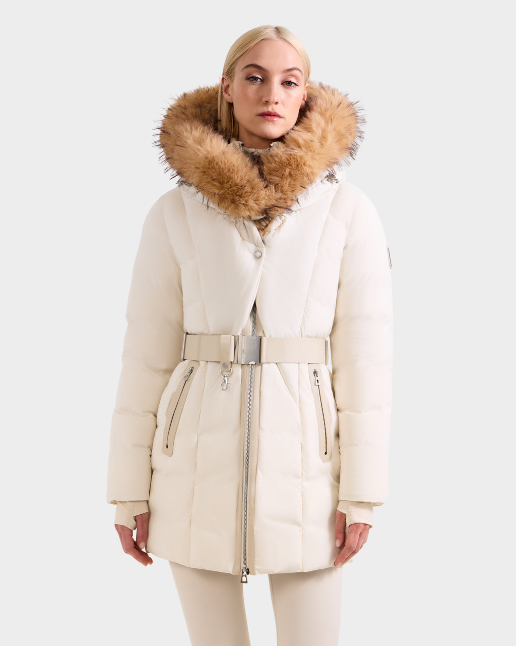 A woman stands facing forward in the Rudsak MODA FF - 8125969 Vanilla, a women’s fitted down puffer with a belted waist and large faux fur hood, set against a plain background.
