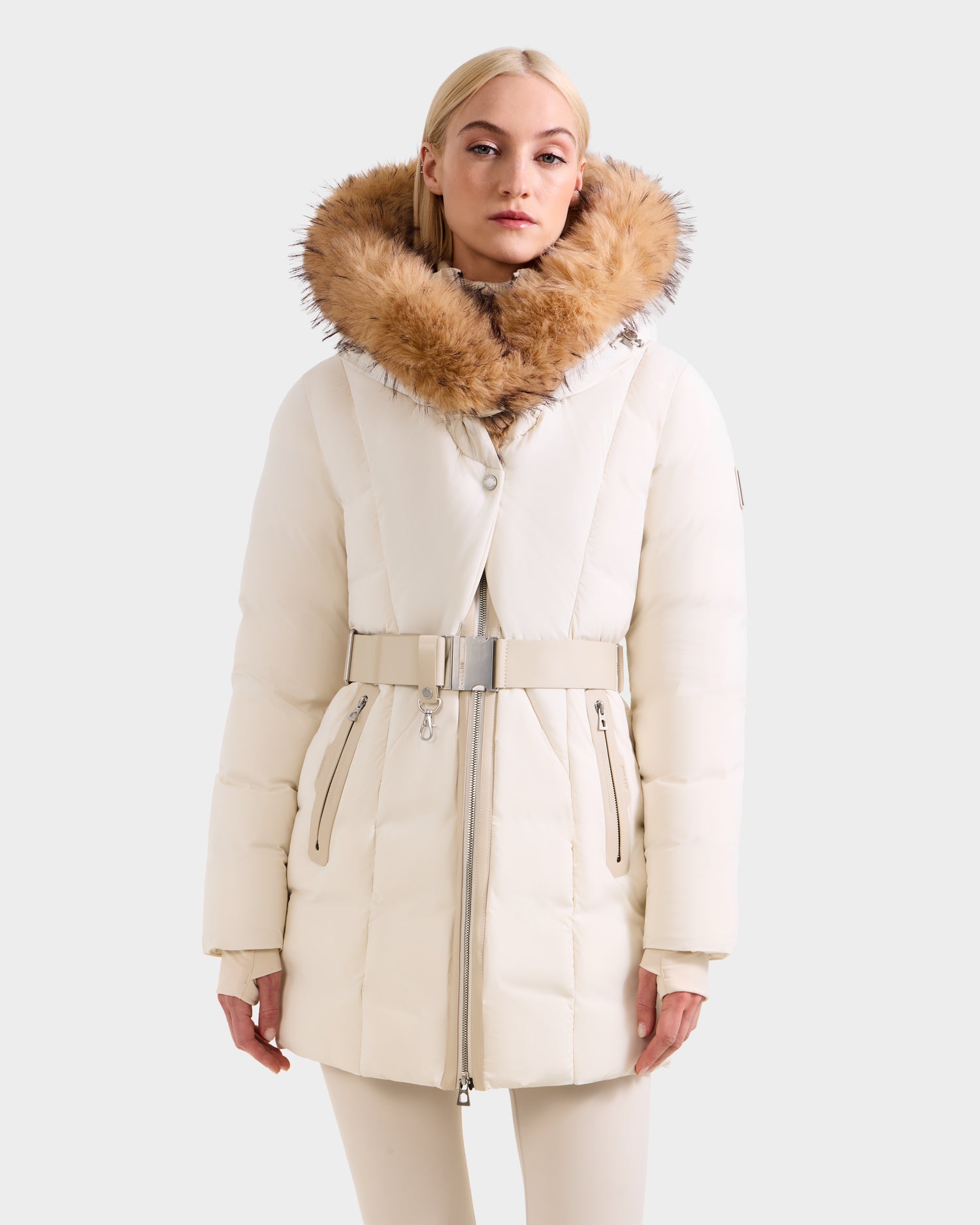 A woman stands facing forward in the Rudsak MODA FF - 8125969 Vanilla, a women’s fitted down puffer with a belted waist and large faux fur hood, set against a plain background.