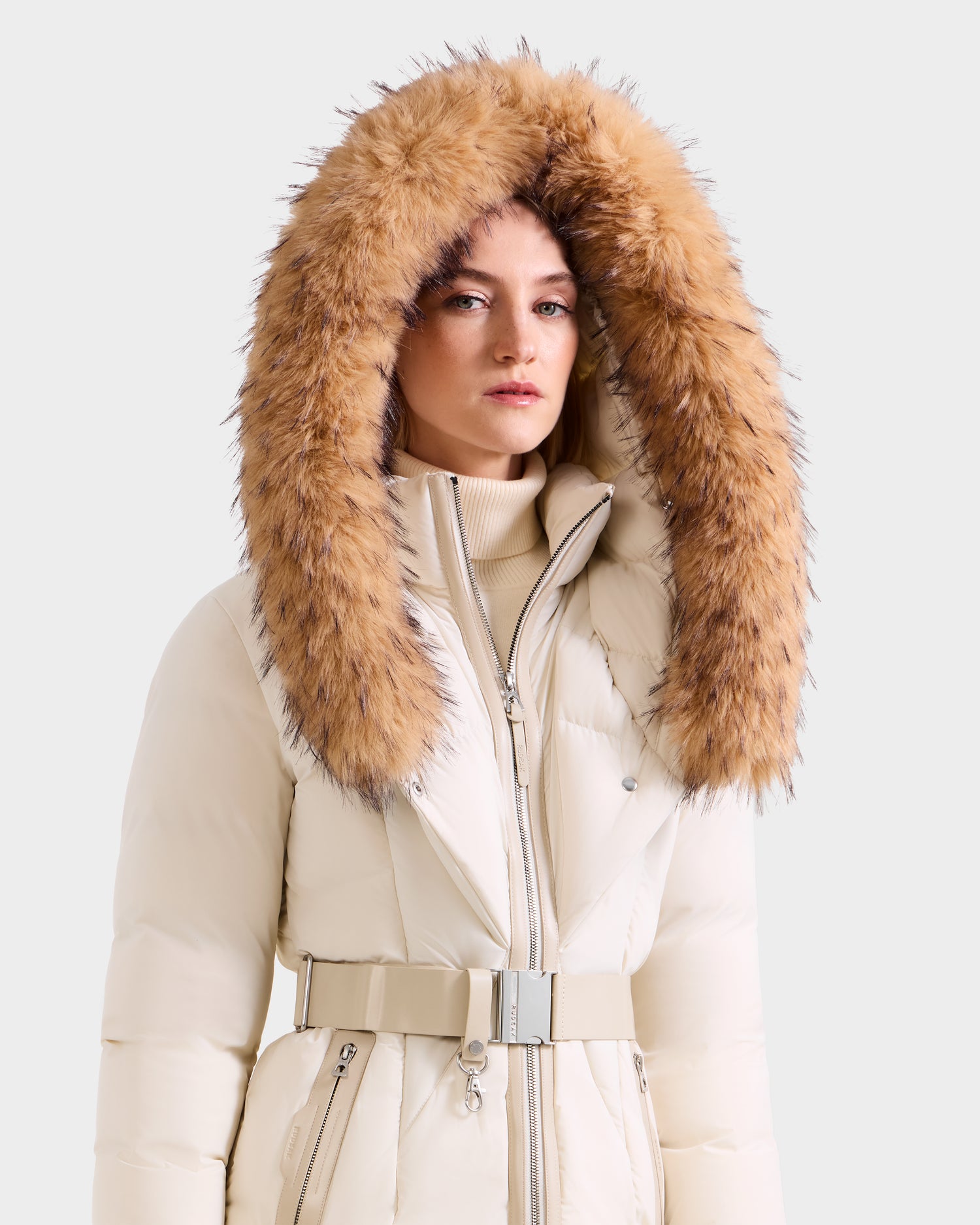 A woman wears the Rudsak MODA FF - 8125969 Vanilla fitted down puffer coat with a large, fluffy faux fur hood in brown and black tones, standing against a plain background and looking at the camera with a neutral expression.