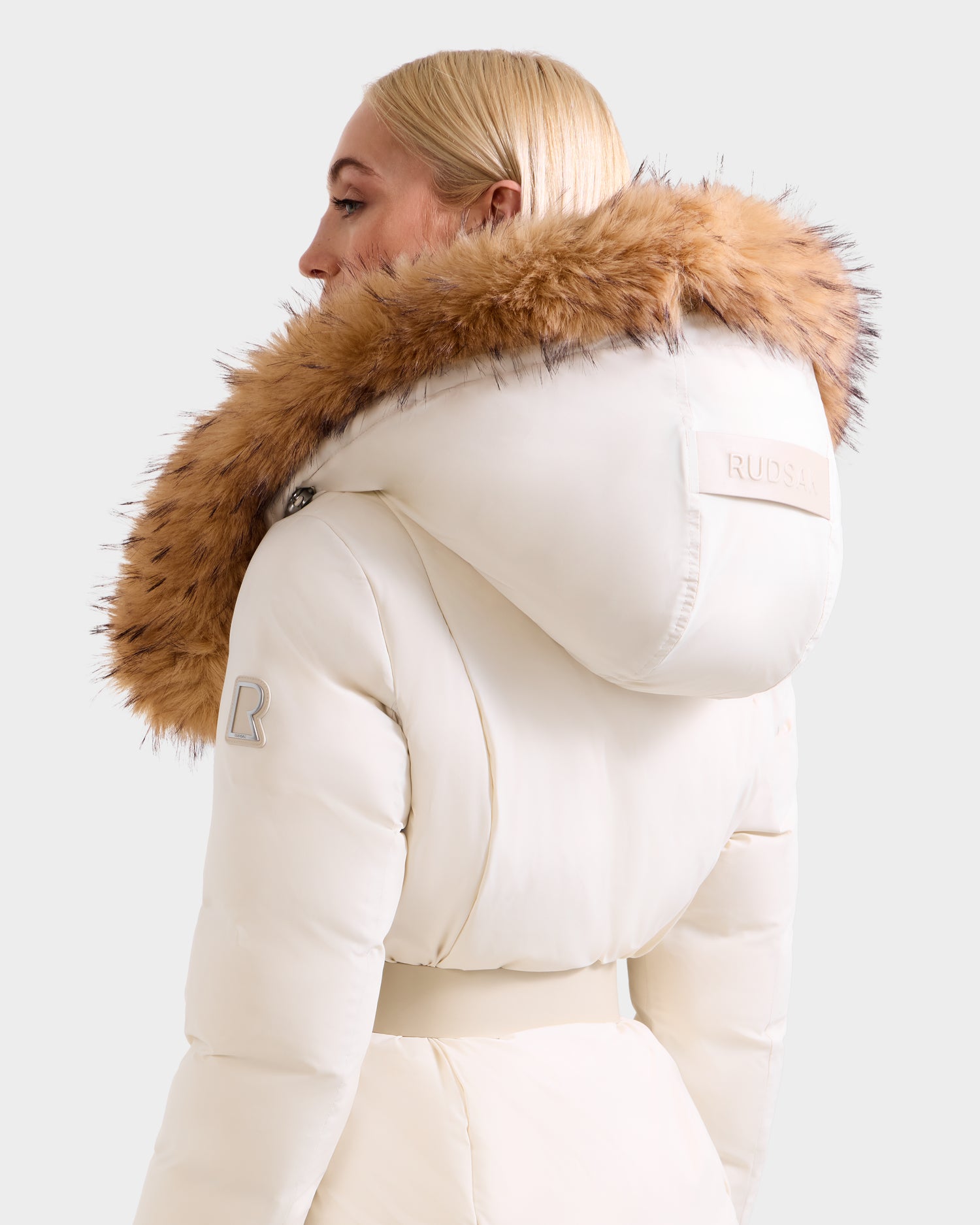 A woman with straight blonde hair wears the Rudsak MODA FF - 8125969 Vanilla fitted down puffer with faux fur hood, seen from the back looking over her left shoulder.