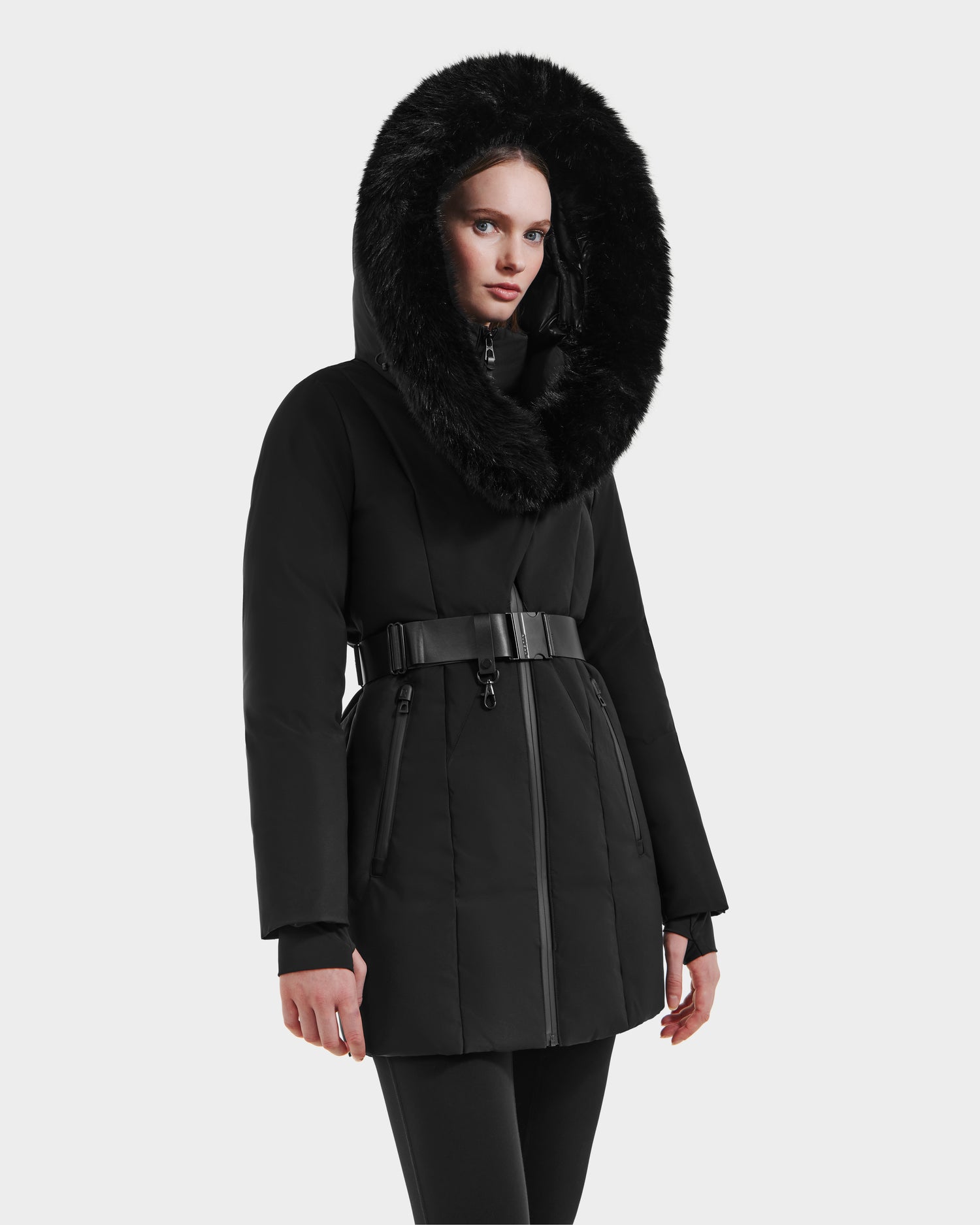 A woman models the Rudsak MODA M - L125555 Black winter coat, featuring a belted design and large fur-trimmed hood, standing against a plain light gray background with her hands relaxed by her sides.