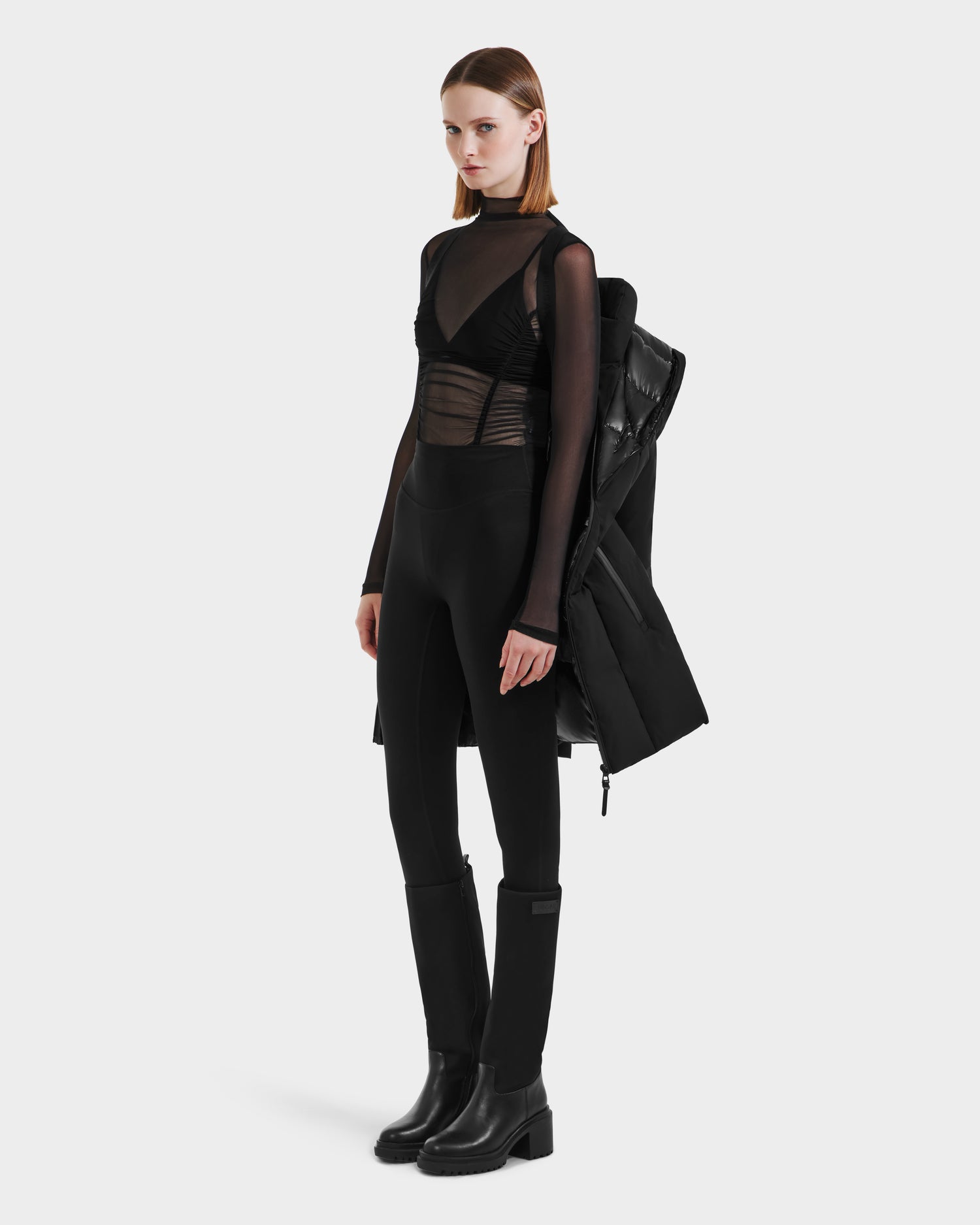 A woman with straight, shoulder-length hair wears the Rudsak MODA M - L125555 Black sheer top, black fitted pants with a vegan leather belt, and black boots, holding a long black coat, standing against a plain white background.