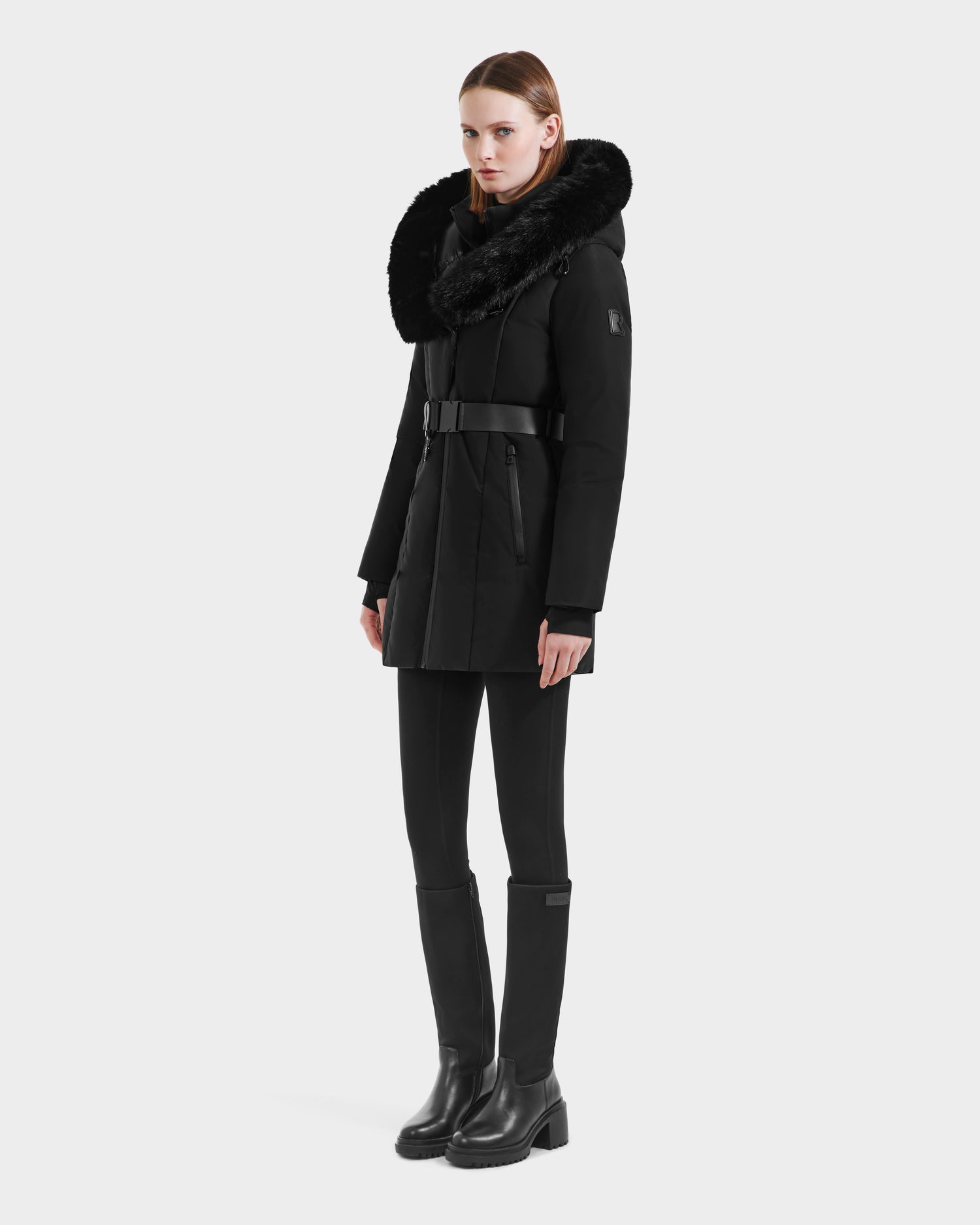 A woman wears the Rudsak MODA M - L125555 Black belted winter coat with a large faux fur hood, styled with black leggings and knee-high boots, standing against a plain white background.