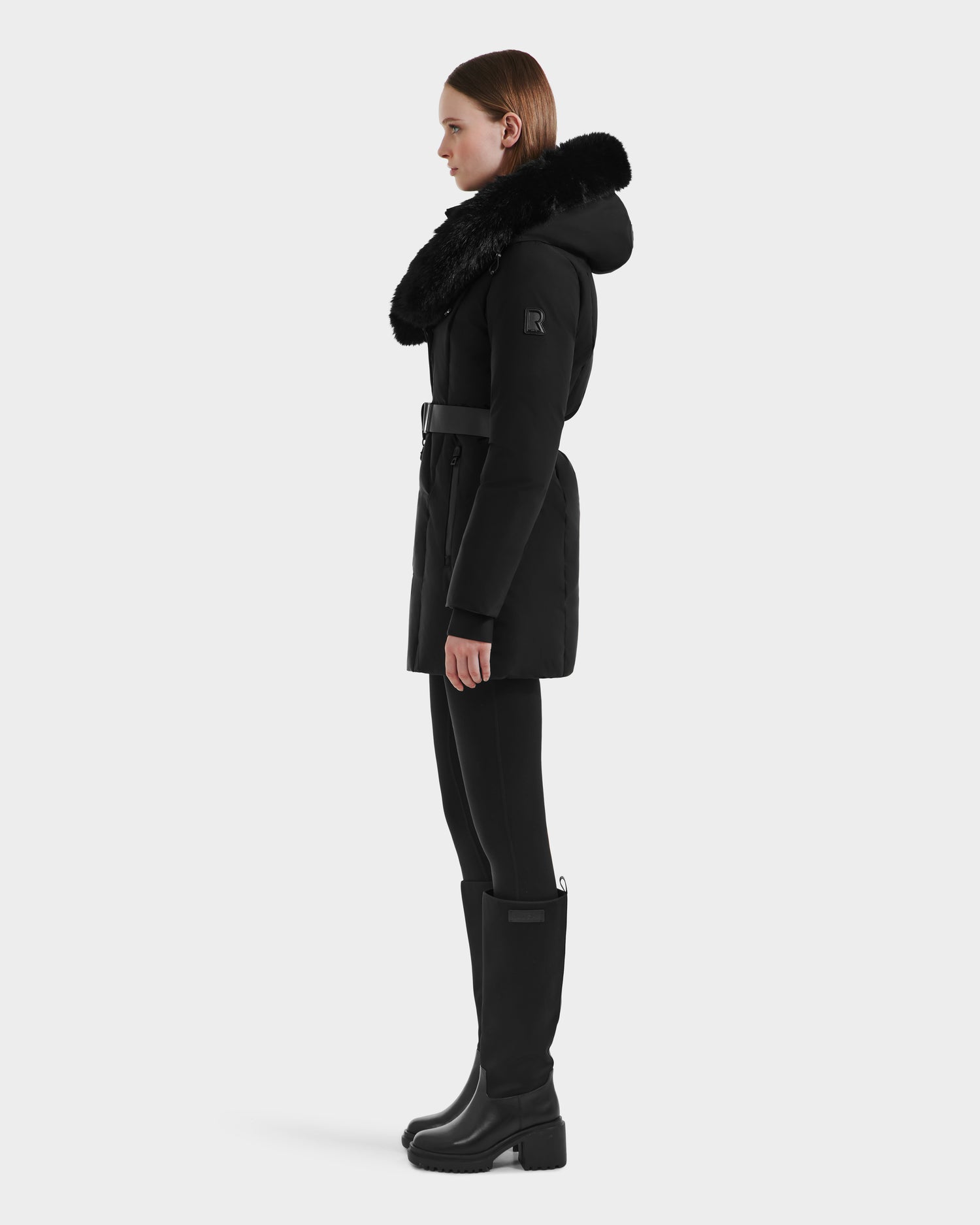 A woman in profile wears the Rudsak MODA M - L125555 Black winter coat with a fur-trimmed hood and vegan leather belt, styled with black pants and knee-high boots against a plain light background.