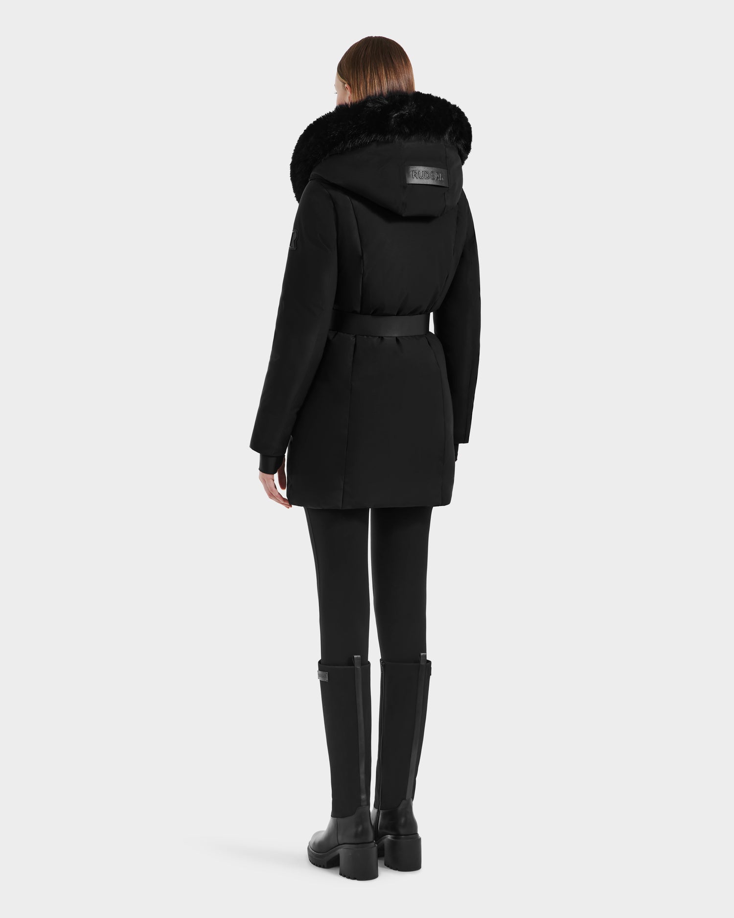 A person in the Rudsak MODA M - L125555 Black hooded winter coat with fur trim, paired with black leggings and knee-high boots, stands facing away from the camera against a plain light background.