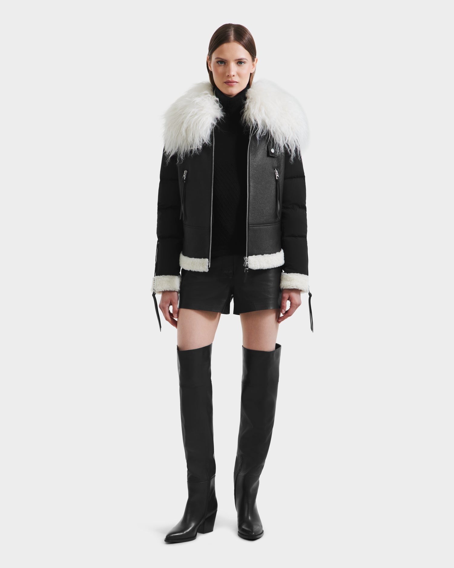 A woman faces forward against a plain white background, wearing the MONKA - 8125629 Black women’s shearling jacket by Rudsak with a large Mongolian collar and cuffs, styled with a black turtleneck, leather shorts, and knee-high boots.