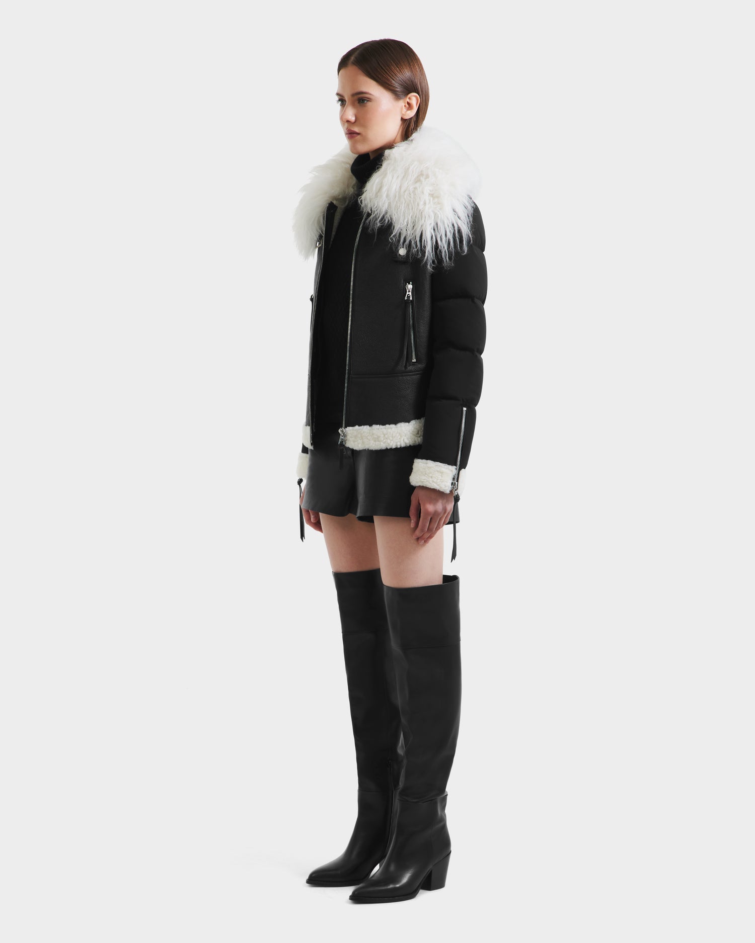A woman stands sideways against a white background, wearing the Rudsak MONKA - 8125629 Black women’s shearling jacket with a bold Mongolian collar, styled with black shorts and over-the-knee boots.