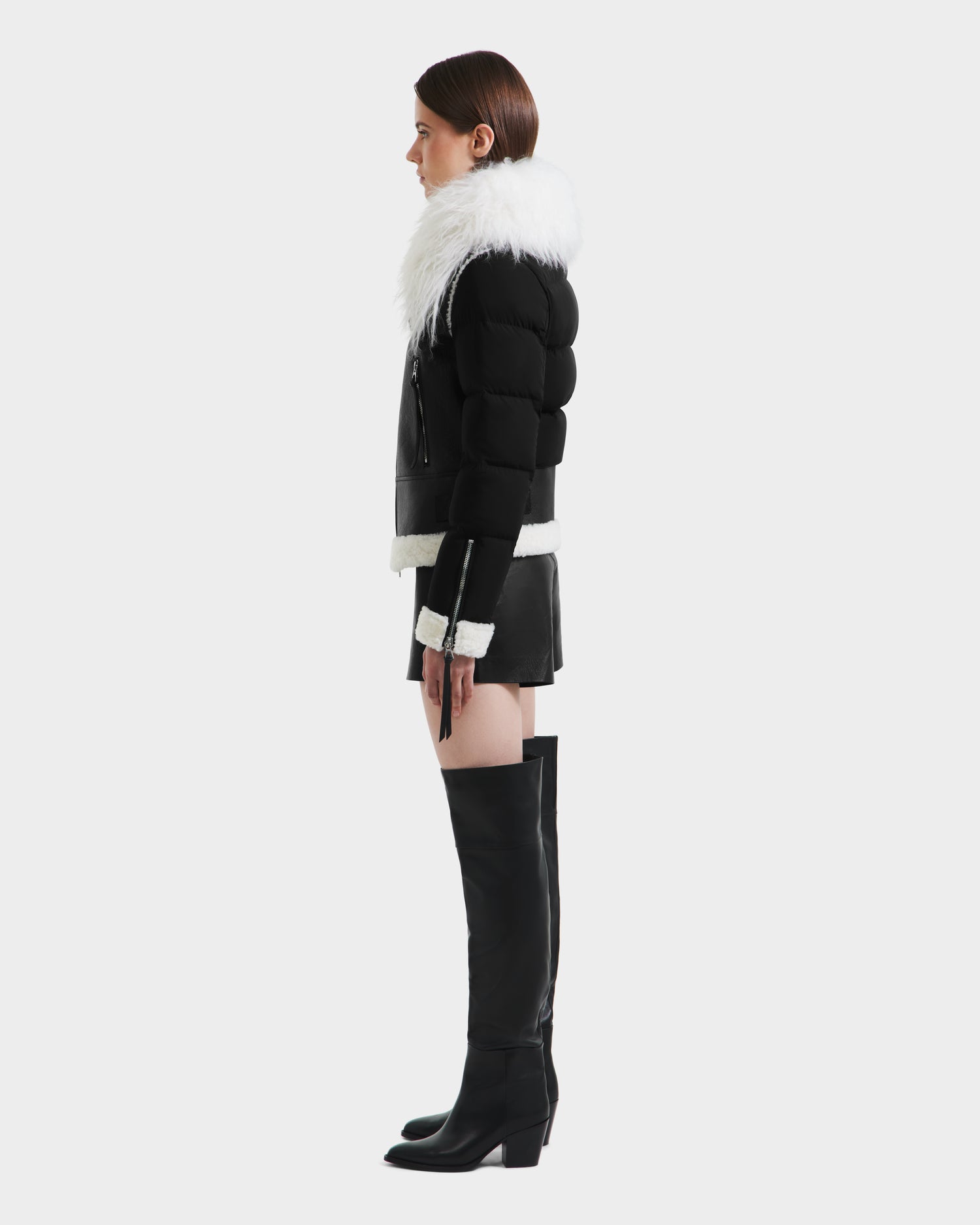 A woman stands in profile wearing the Rudsak MONKA - 8125629 Black women's shearling jacket with a large white Mongolian collar, paired with a black mini skirt and tall knee-high boots against a plain light gray background.