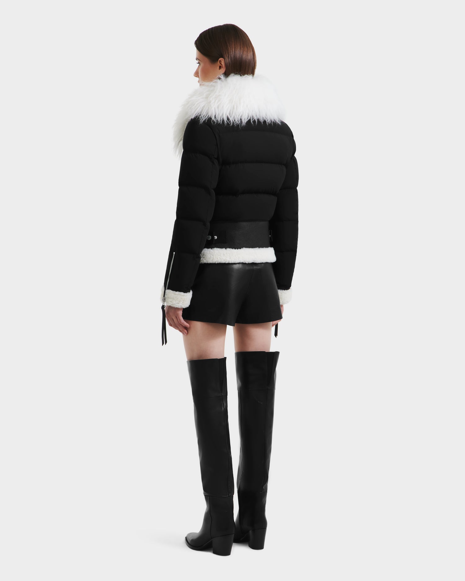 A woman faces away, wearing the Rudsak MONKA - 8125629 Black lightweight down jacket with a Mongolian shearling collar, paired with black shorts and knee-high boots against a plain light background.