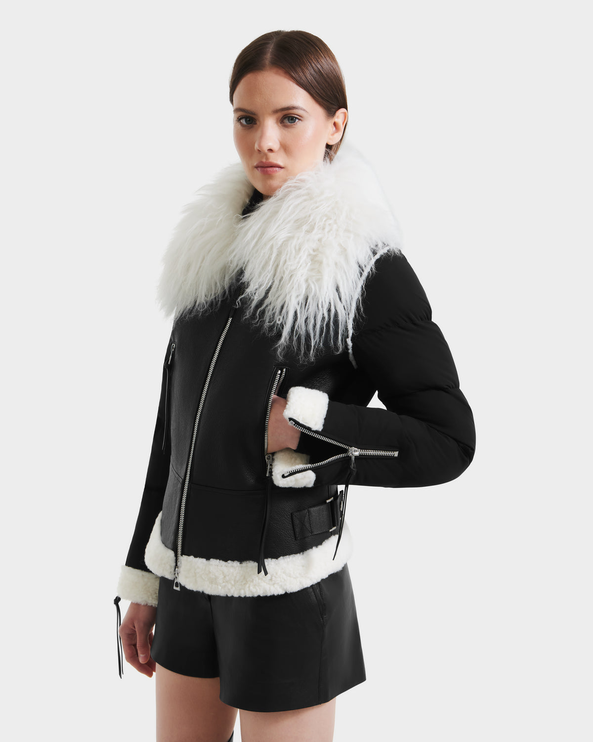 A woman stands against a plain white background wearing the Rudsak MONKA - 8125629 Black women’s shearling jacket with a white faux fur collar and trim, paired with black shorts.