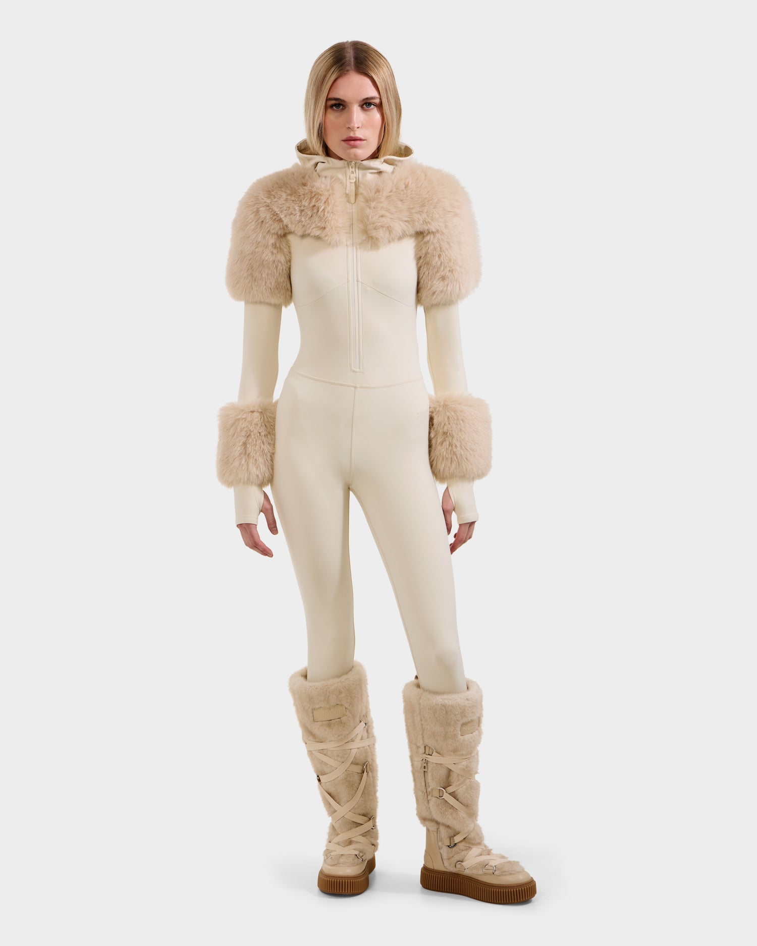 A woman with straight blonde hair and a neutral expression faces forward in the Rudsak MYLA - 8525671 Vanilla cream bodysuit, featuring faux fur trim on the shoulders, cuffs, and knee-high boots. Set against a plain white background.