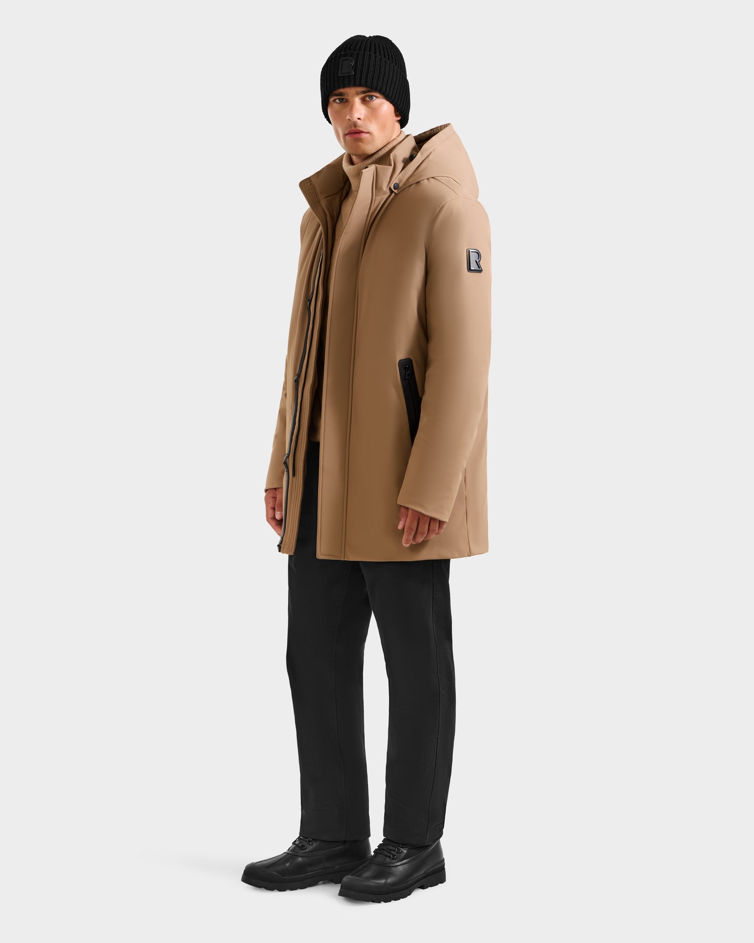 A man stands against a plain white background, looking slightly to the side, wearing the Rudsak NATAN - M124519 Sand men's parka, paired with a black beanie, black pants, and black shoes.