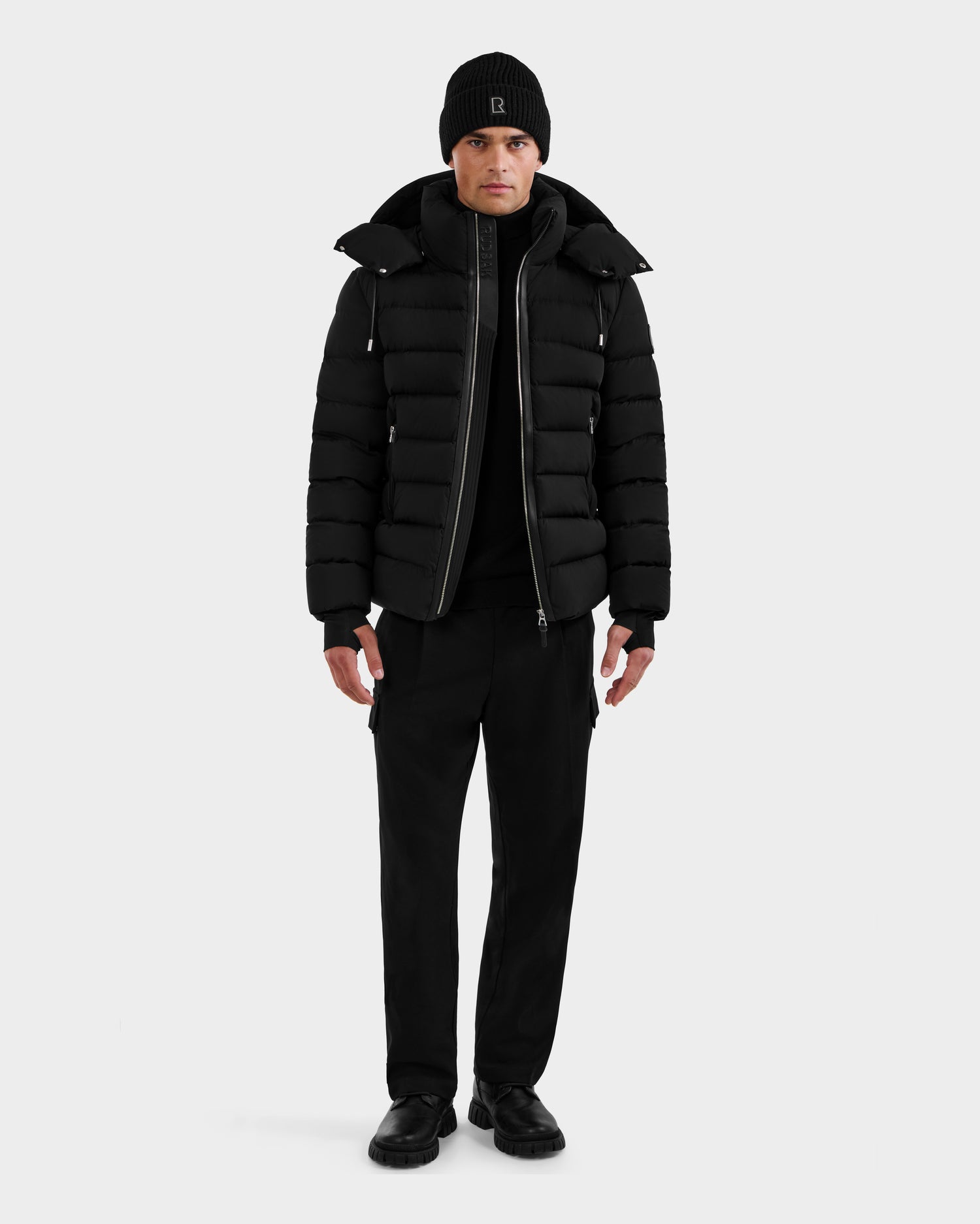 A man faces forward wearing the Rudsak NAVAN - 6125598 Black men's down puffer jacket, paired with a black beanie, pants, shoes, and shirt, set against a plain light background.