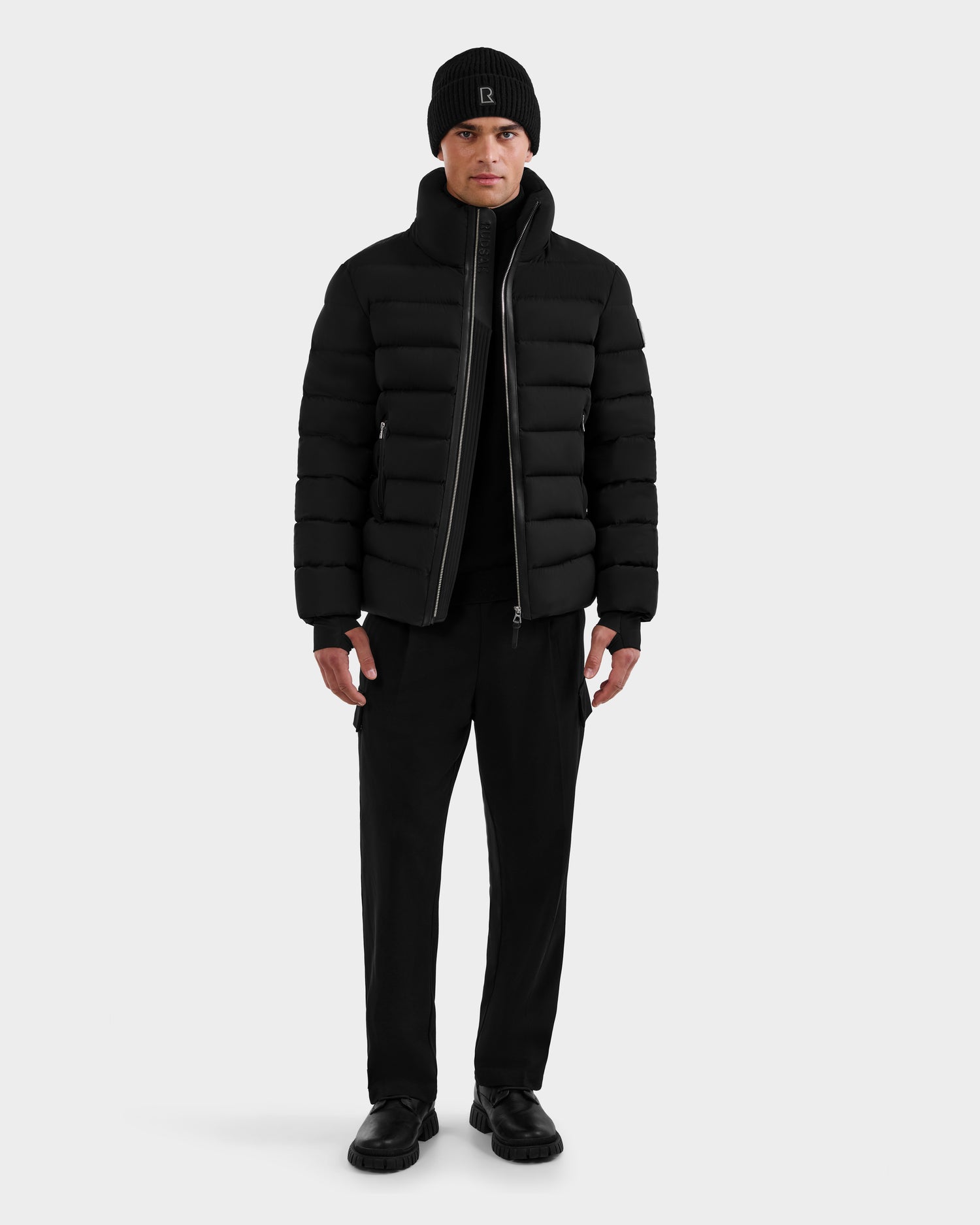 A person faces forward against a white background, wearing the Rudsak NAVAN - 6125598 Black quilted puffer jacket filled with RDS-certified down, paired with a black beanie, black pants, and black shoes.