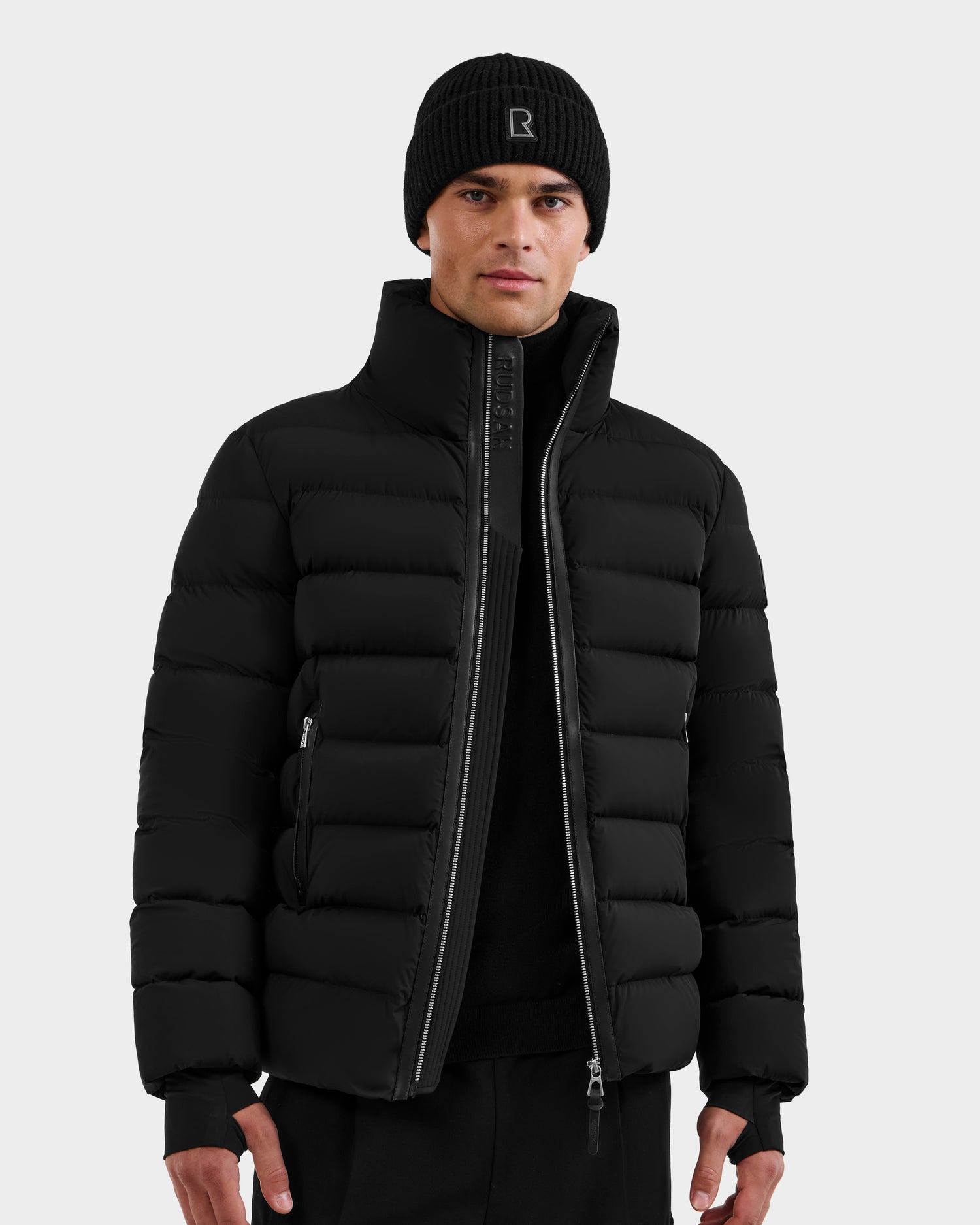 A man stands facing forward with a neutral expression, wearing the Rudsak NAVAN - 6125598 Black men's down puffer jacket, black knit beanie, and black pants against a plain light gray background.