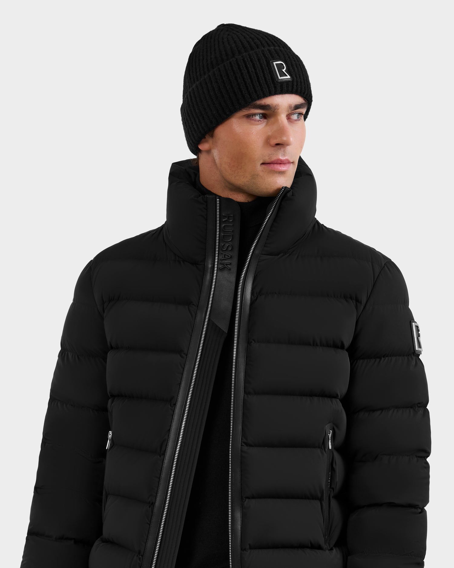A man in a Rudsak NAVAN - 6125598 Black RDS-certified down puffer jacket with a high collar and a black knit beanie stands against a plain light background.