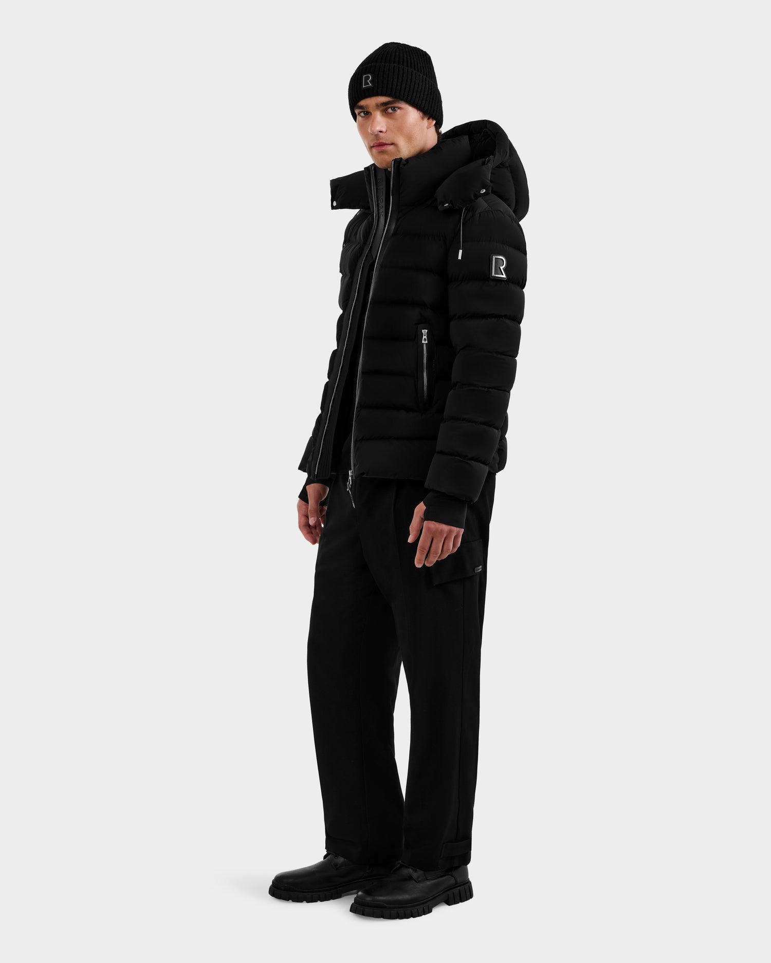 A person wearing the Rudsak NAVAN - 6125598 Black RDS-certified down puffer jacket, along with a black beanie, pants, and shoes, stands turned slightly to the side against a plain light background.