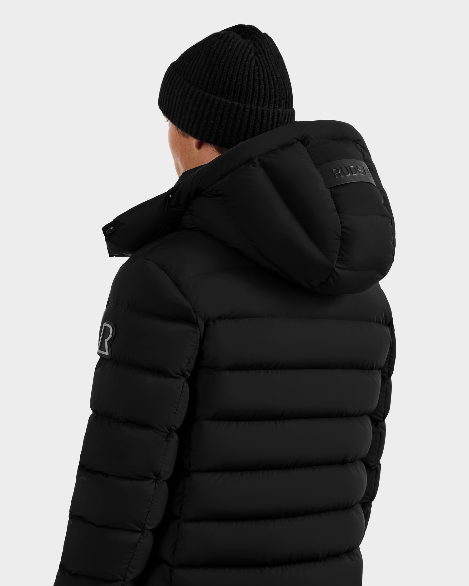 A person is seen from behind wearing the Rudsak NAVAN - 6125598 Black men's down puffer with a hood, shown against a plain light background. The quilted jacket features a sleeve patch and a logo on the back of the hood.