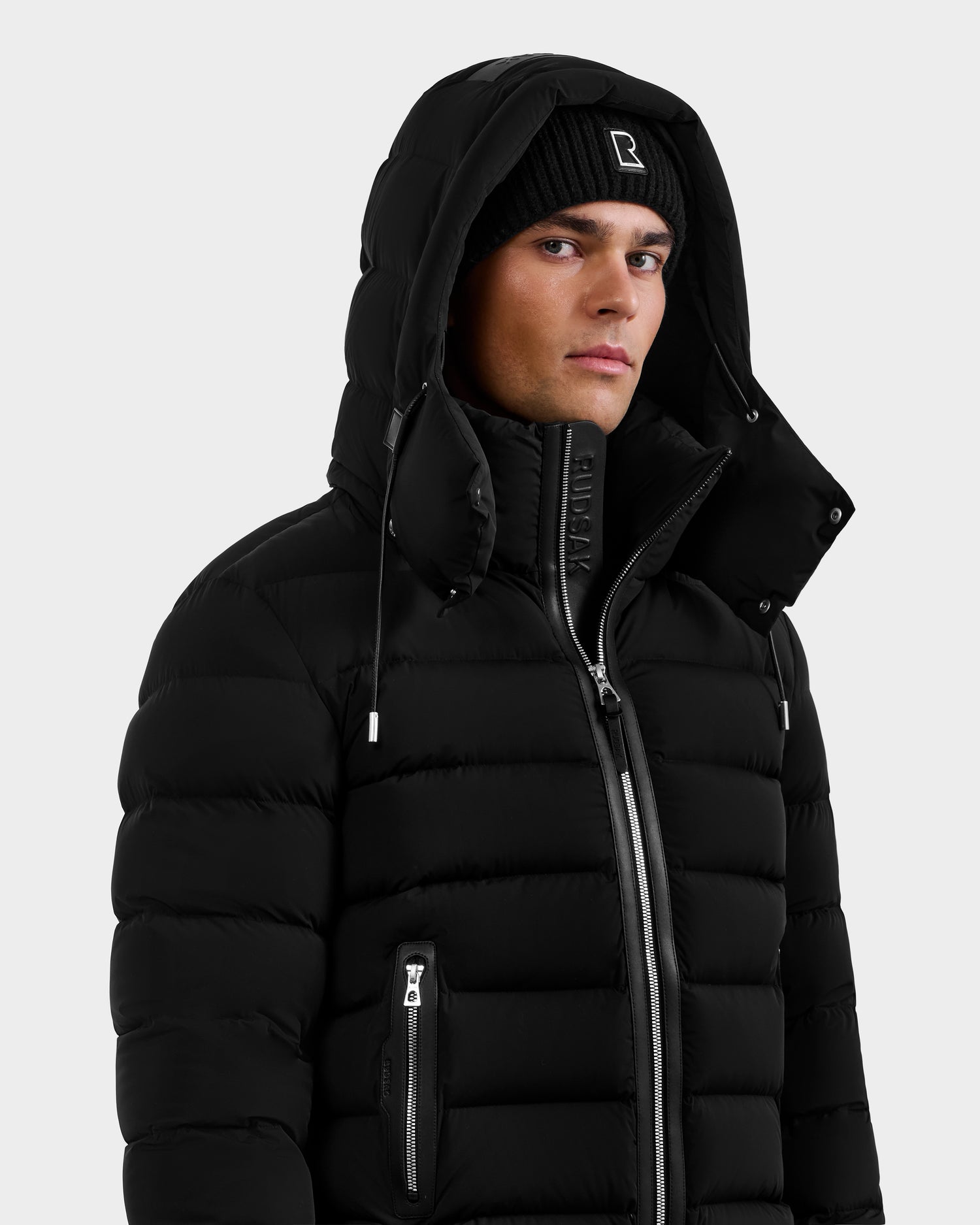 A man in the Rudsak NAVAN - 6125598 Black men's down puffer jacket with a hood and a black beanie stands against a plain light background, gazing slightly to the side with a neutral expression.