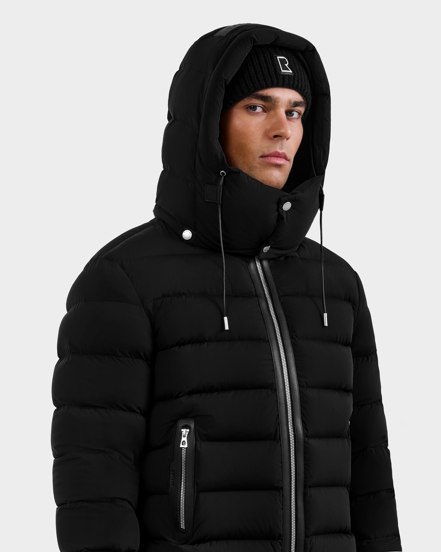 A man wearing the Rudsak NAVAN - 6125598 Black puffer jacket with a hood up and a black beanie looks to the side against a plain light gray background.