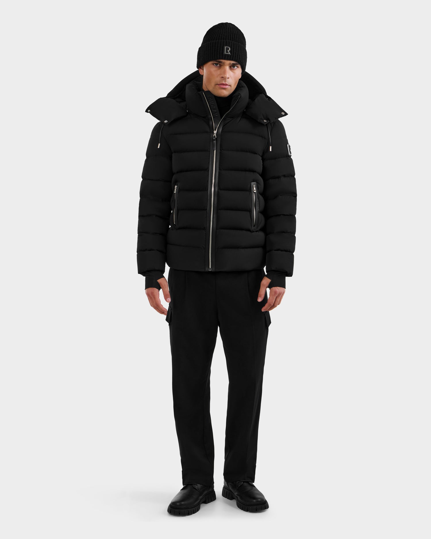 A person faces forward against a white background, wearing the Rudsak NAVAN - 6125598 Black quilted puffer jacket, along with a black beanie, black pants, and black shoes.