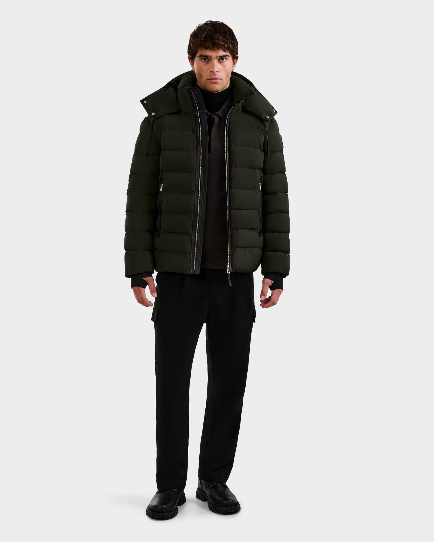 A person faces forward, wearing the Rudsak NAVAN - 6125598 Dark Olive men’s down puffer jacket with black pants and black shoes against a plain white background.