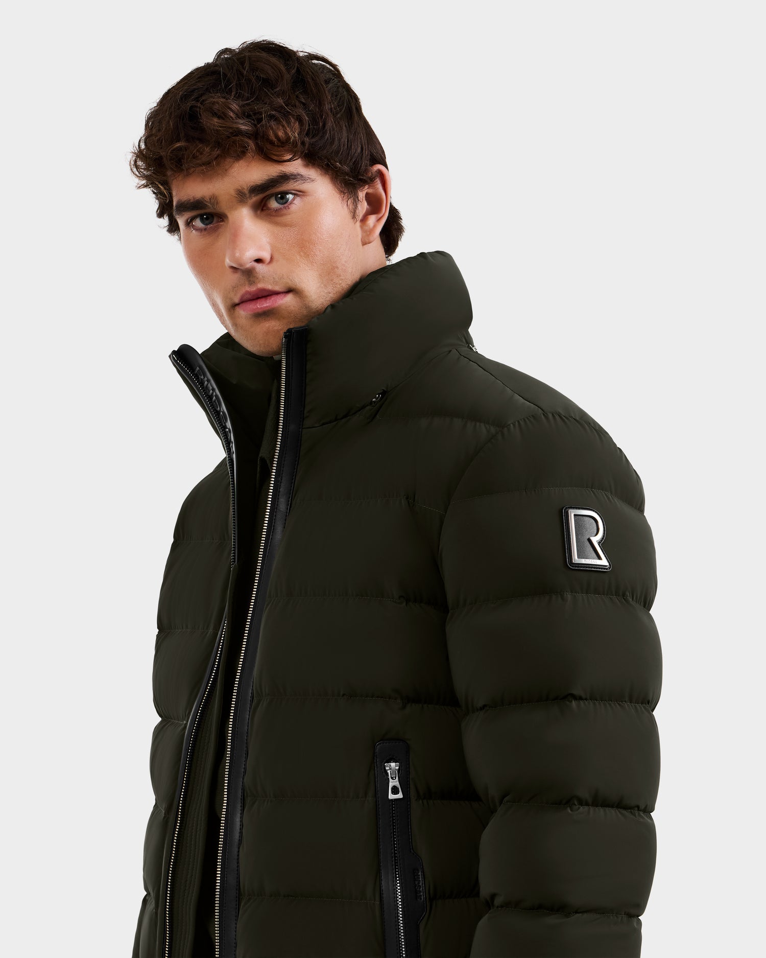 A young man with curly brown hair wears the Rudsak NAVAN - 6125598 dark olive puffer jacket featuring a high collar, zippered pockets, and an "R" patch on the left sleeve, standing against a plain light background.