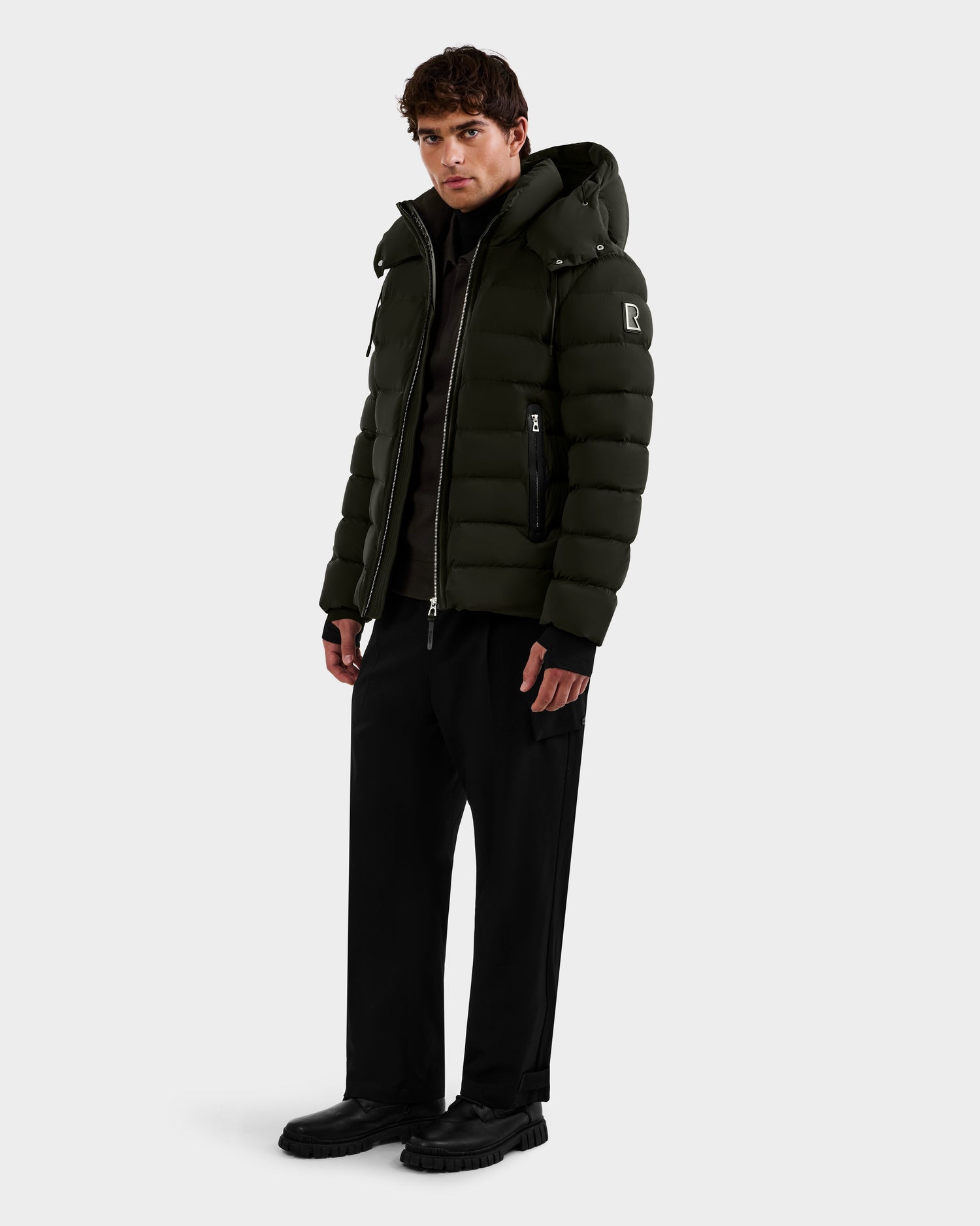 A man with short brown hair and a neutral expression stands against a plain background wearing the Rudsak NAVAN - 6125598 Dark Olive men's quilted puffer jacket with a hood, black wide-leg pants, and black shoes.