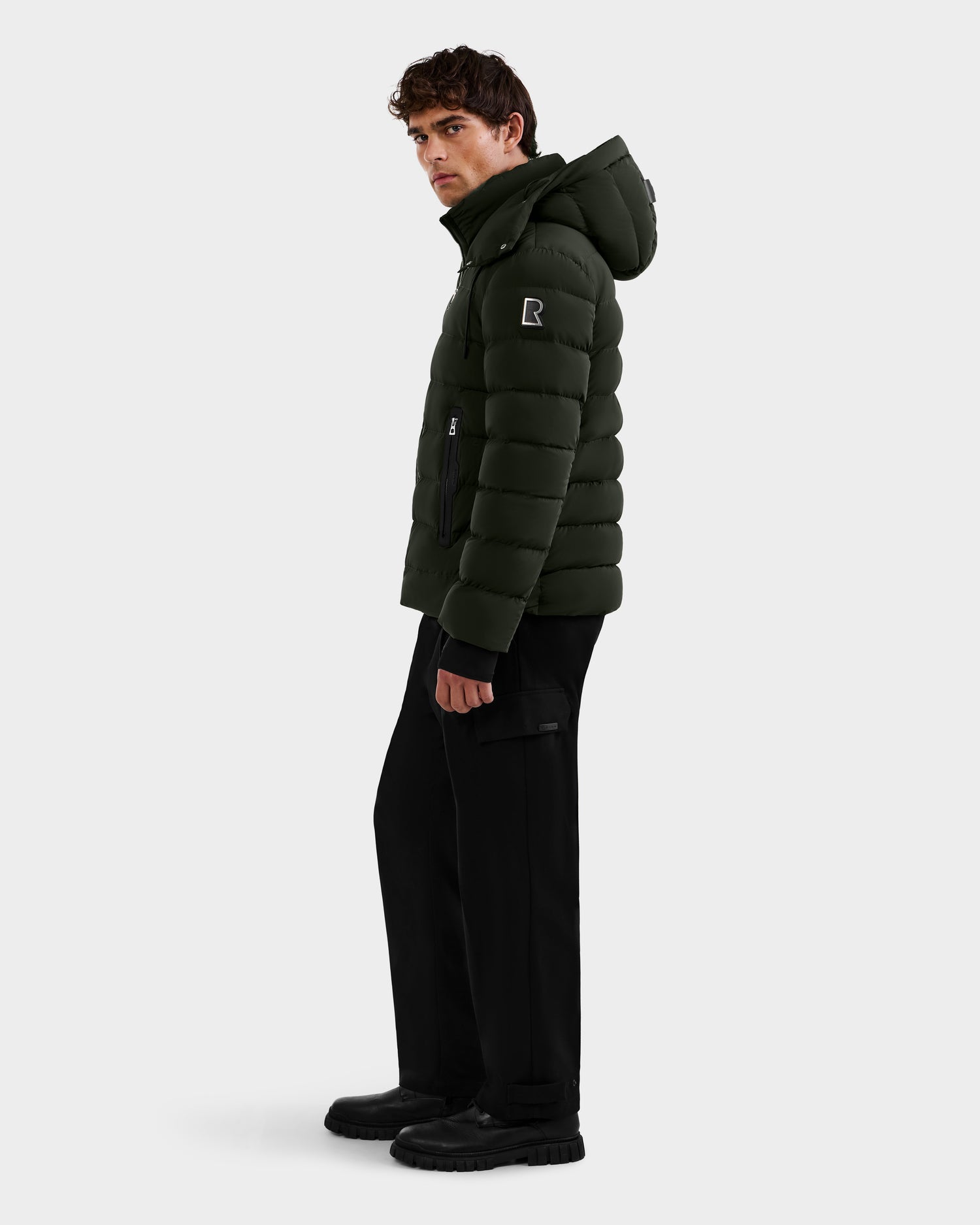 A man wearing the Rudsak NAVAN - 6125598 Dark Olive quilted puffer jacket with hood and black pants stands sideways, glancing over his shoulder, set against a plain light background.
