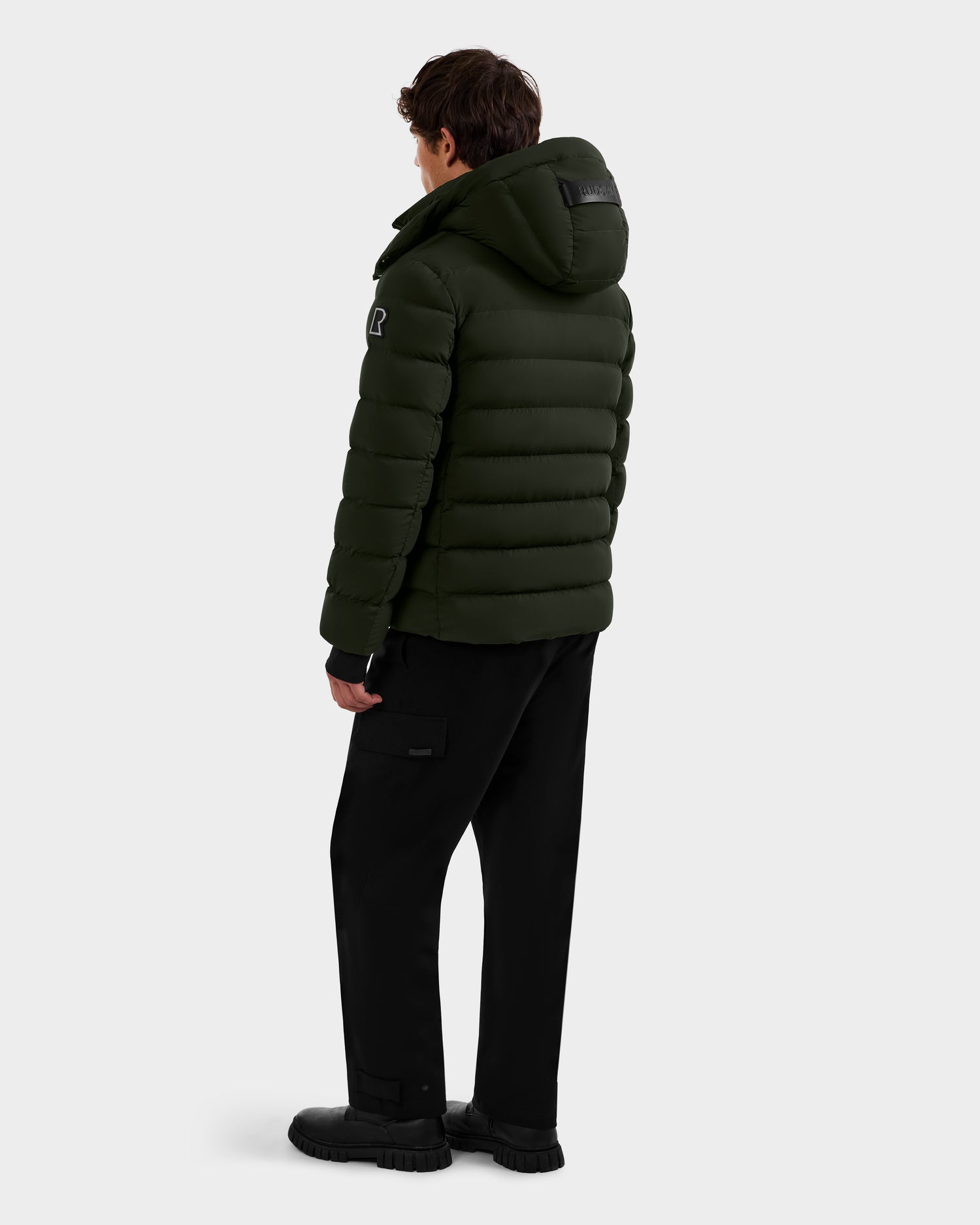 A person stands facing away, wearing the Rudsak NAVAN - 6125598 Dark Olive men’s down puffer jacket with a hood and black pants. The light background highlights its quilted design and RDS-certified down.