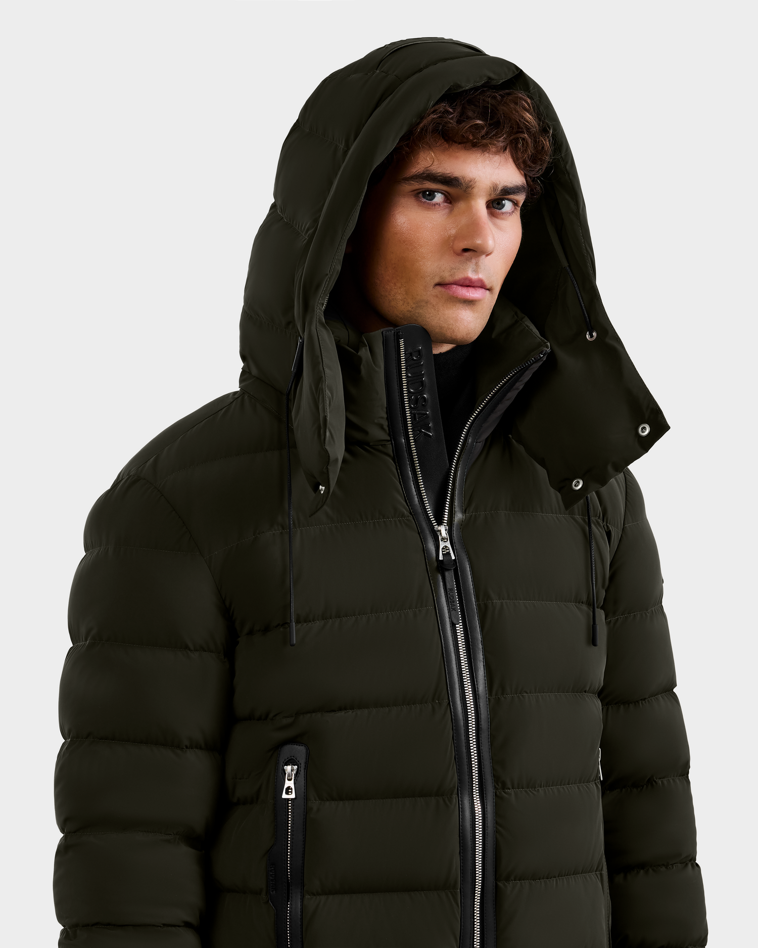 A young man wearing the Rudsak NAVAN - 6125598 Dark Olive RDS-certified down puffer jacket with hood, partially zipped over a black layer, stands against a plain light background.