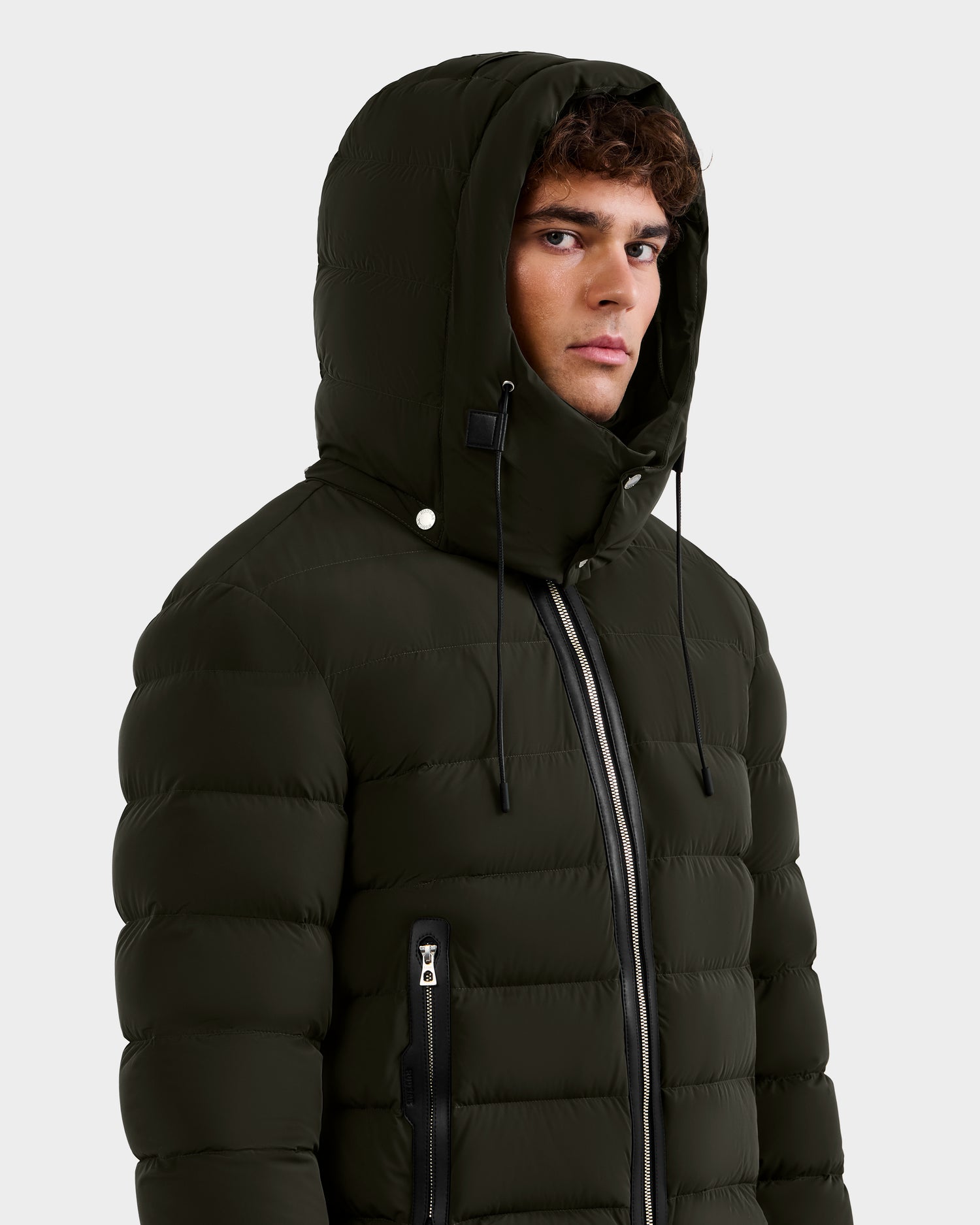 A young man stands against a plain light background, looking slightly to the side, wearing the Rudsak NAVAN - 6125598 down puffer jacket in dark olive with the hood up.
