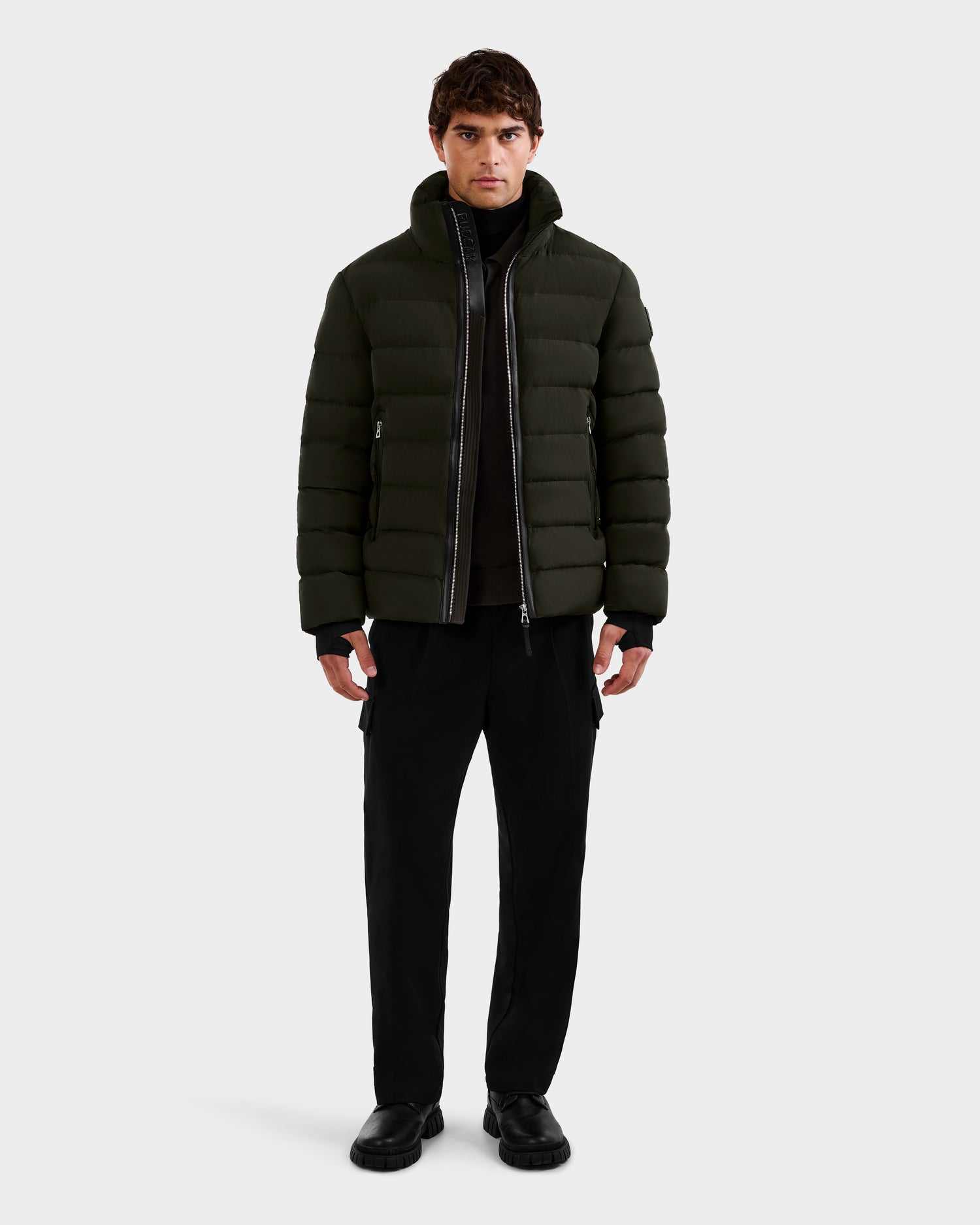 A man stands facing forward in the Rudsak NAVAN - 6125598 Dark Olive quilted puffer, worn over a black shirt and pants with black shoes. This men's down jacket uses RDS-certified down for warmth and sustainability against a plain light background.