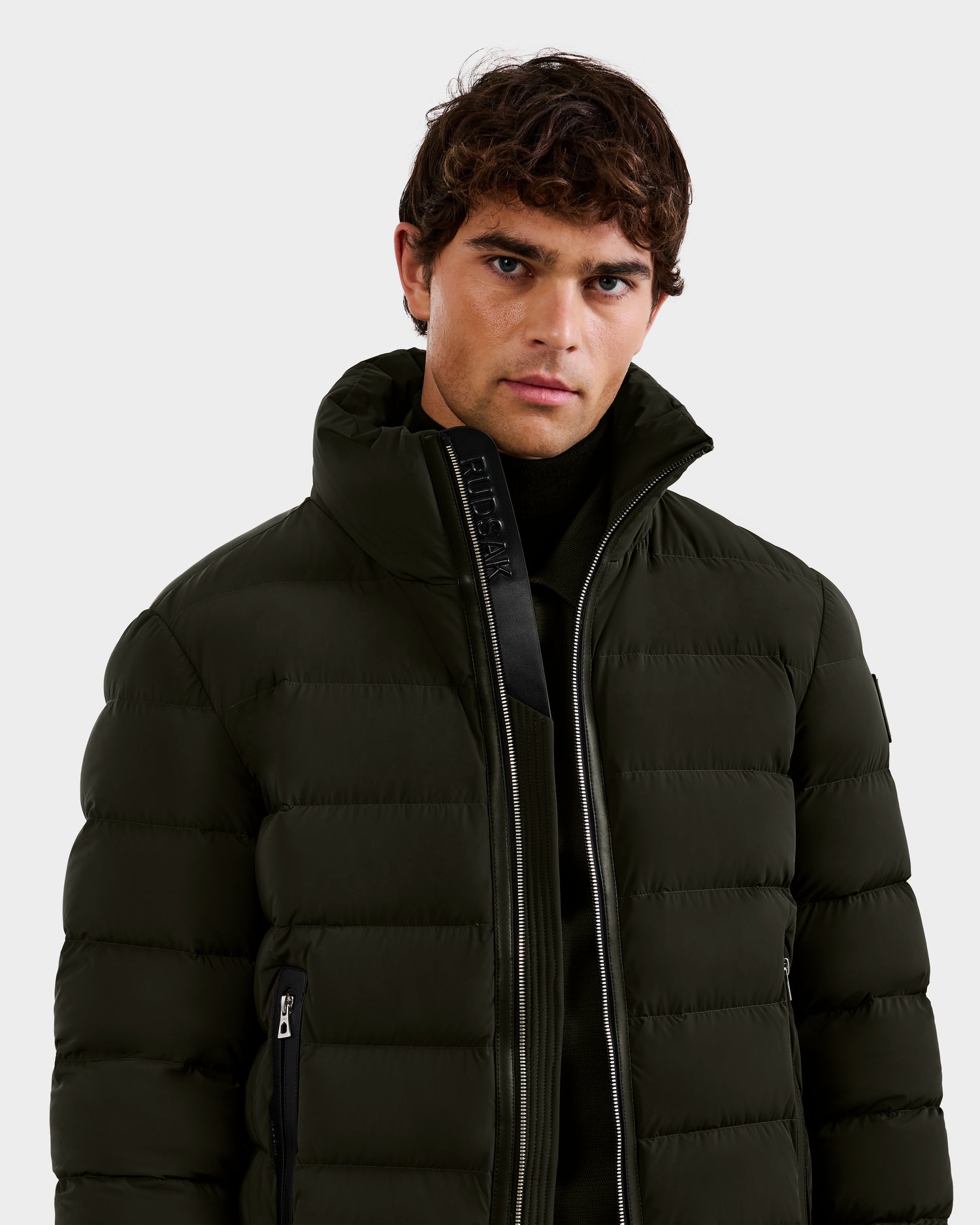 A young man with short, curly brown hair models the Rudsak NAVAN - 6125598 Dark Olive quilted puffer jacket, its high collar partially unzipped to show a black layer beneath, standing against a plain light gray background.