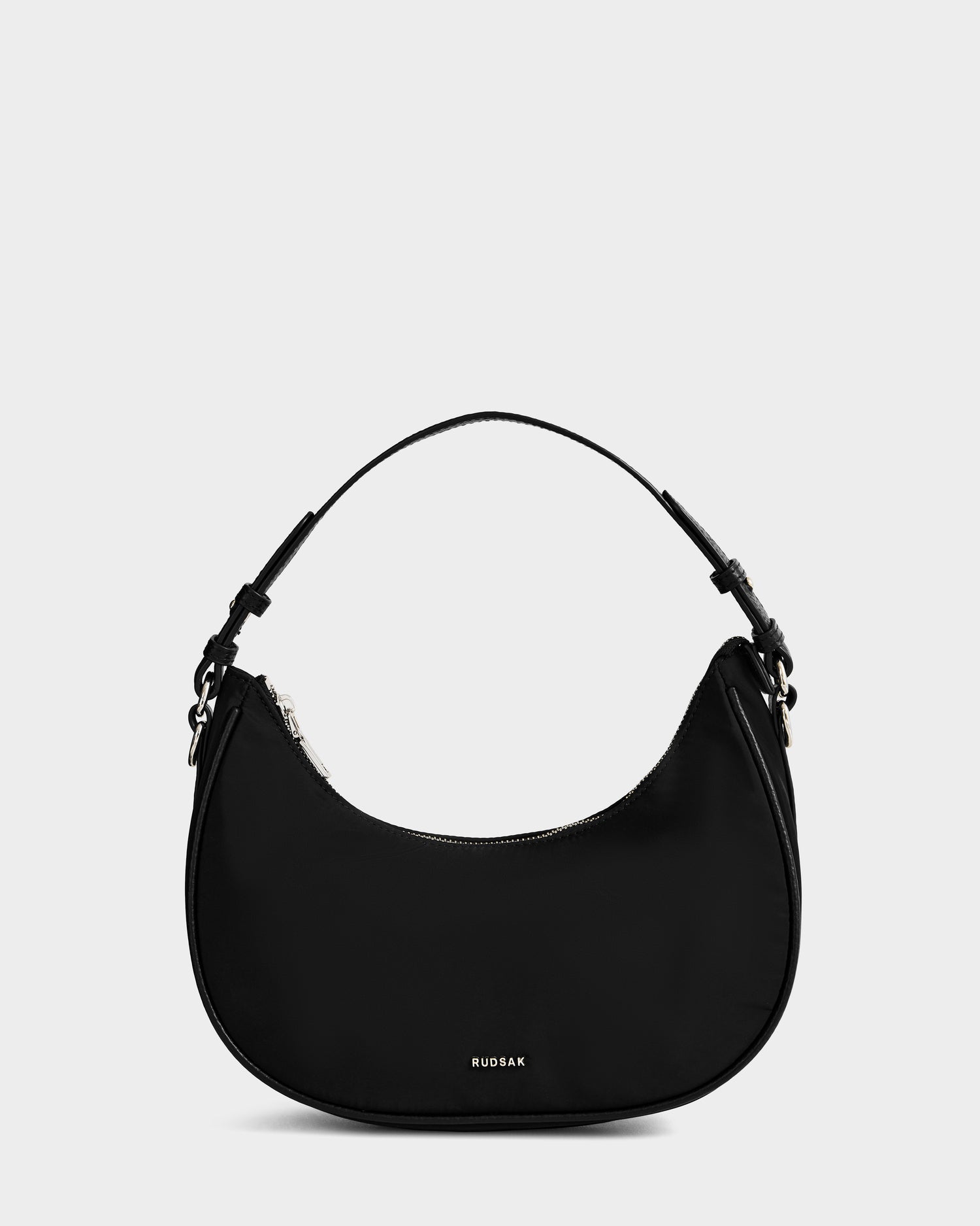 The Rudsak NIKKI - L325512 Black is a women’s crescent shoulder bag with a short strap, zippered top, small silver hardware, and “RUDSAK” in white on the front, displayed against a plain light background.