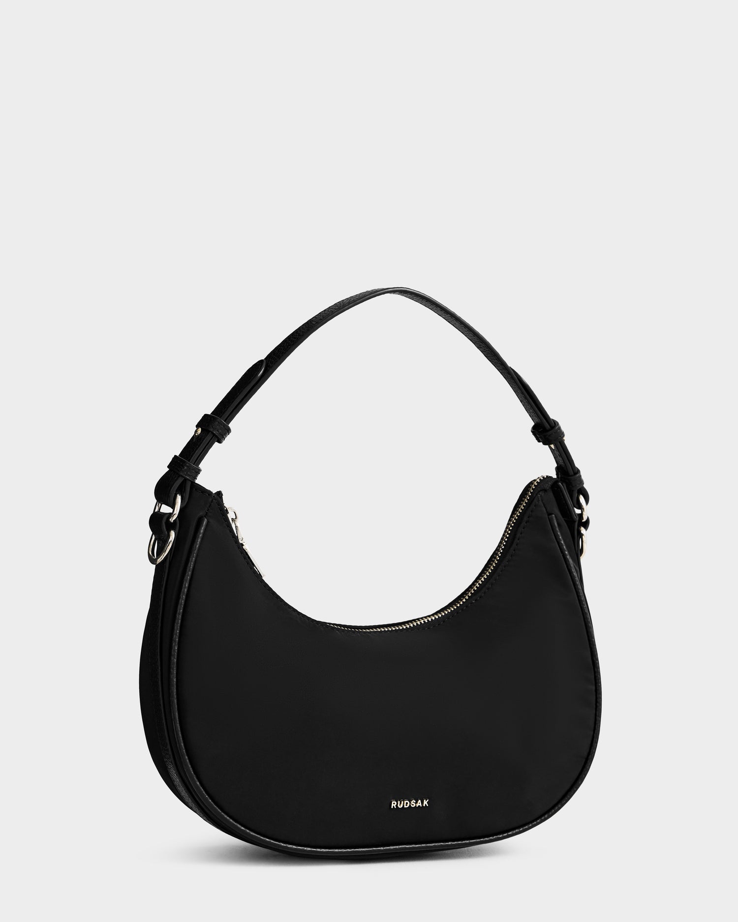 The Rudsak NIKKI - L325512 Black is a women’s black crescent shoulder bag featuring a single strap, gold-tone zipper, and subtle gold RUDSAK logo on the front, shown against a plain light gray background.