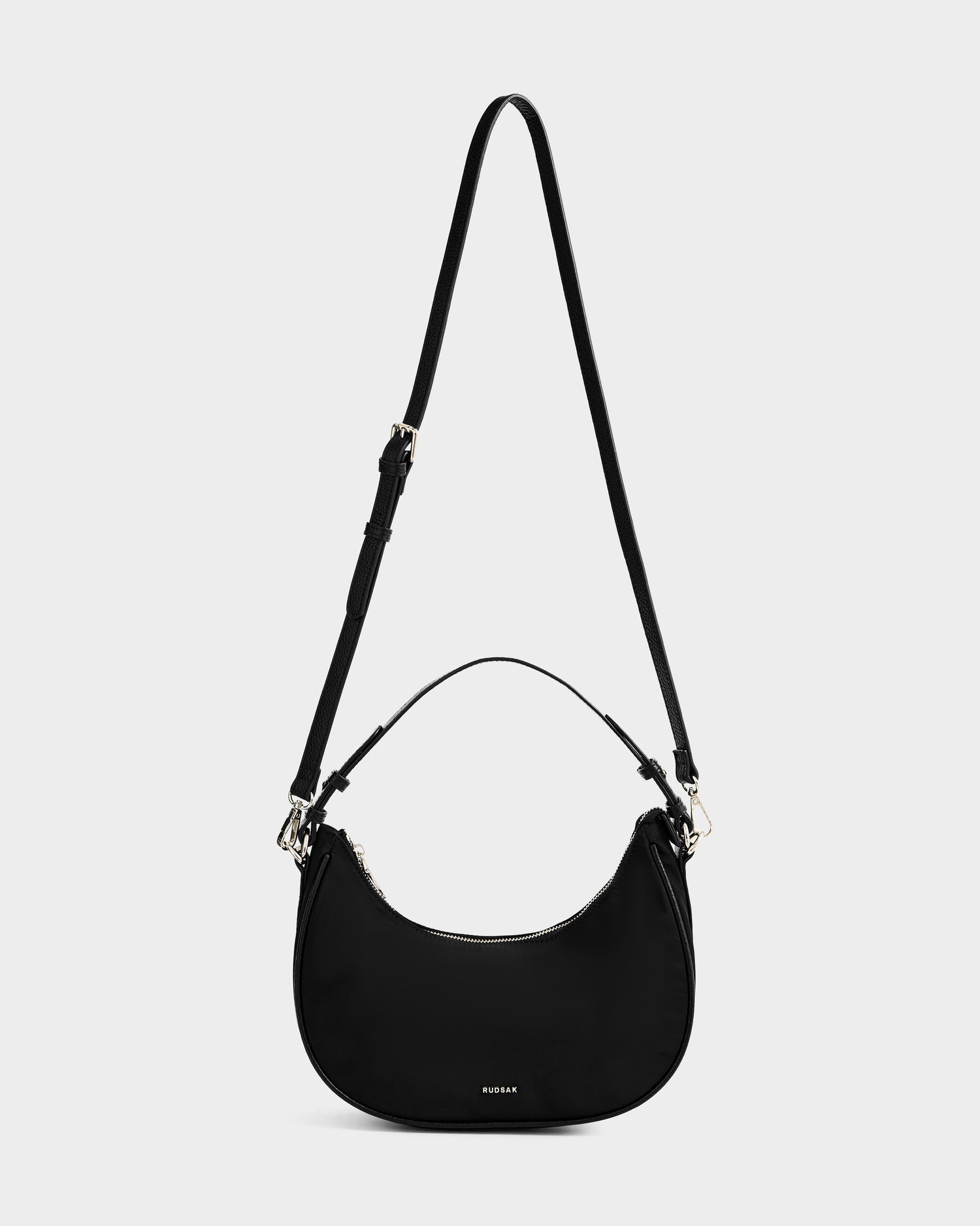 The NIKKI - L325512 Black by Rudsak is a black women’s crescent hobo bag with a short handle, long adjustable crossbody strap, silver hardware, and a small RUMAR logo on the front, shown against a white background.