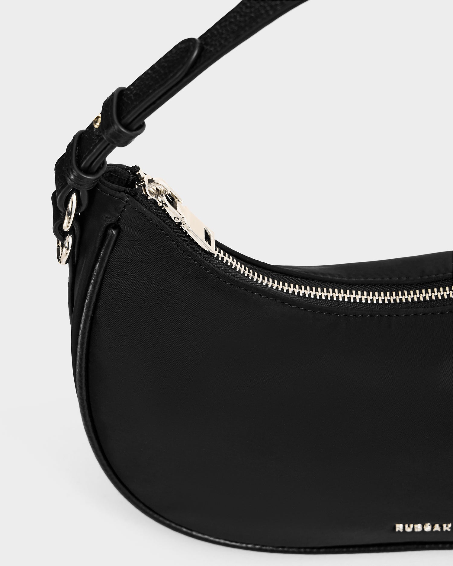 Close-up of the Rudsak NIKKI - L325512 Black women's crescent shoulder bag, featuring a textured strap, silver zipper, and small metallic logo on the bottom right corner, set against a plain light background.