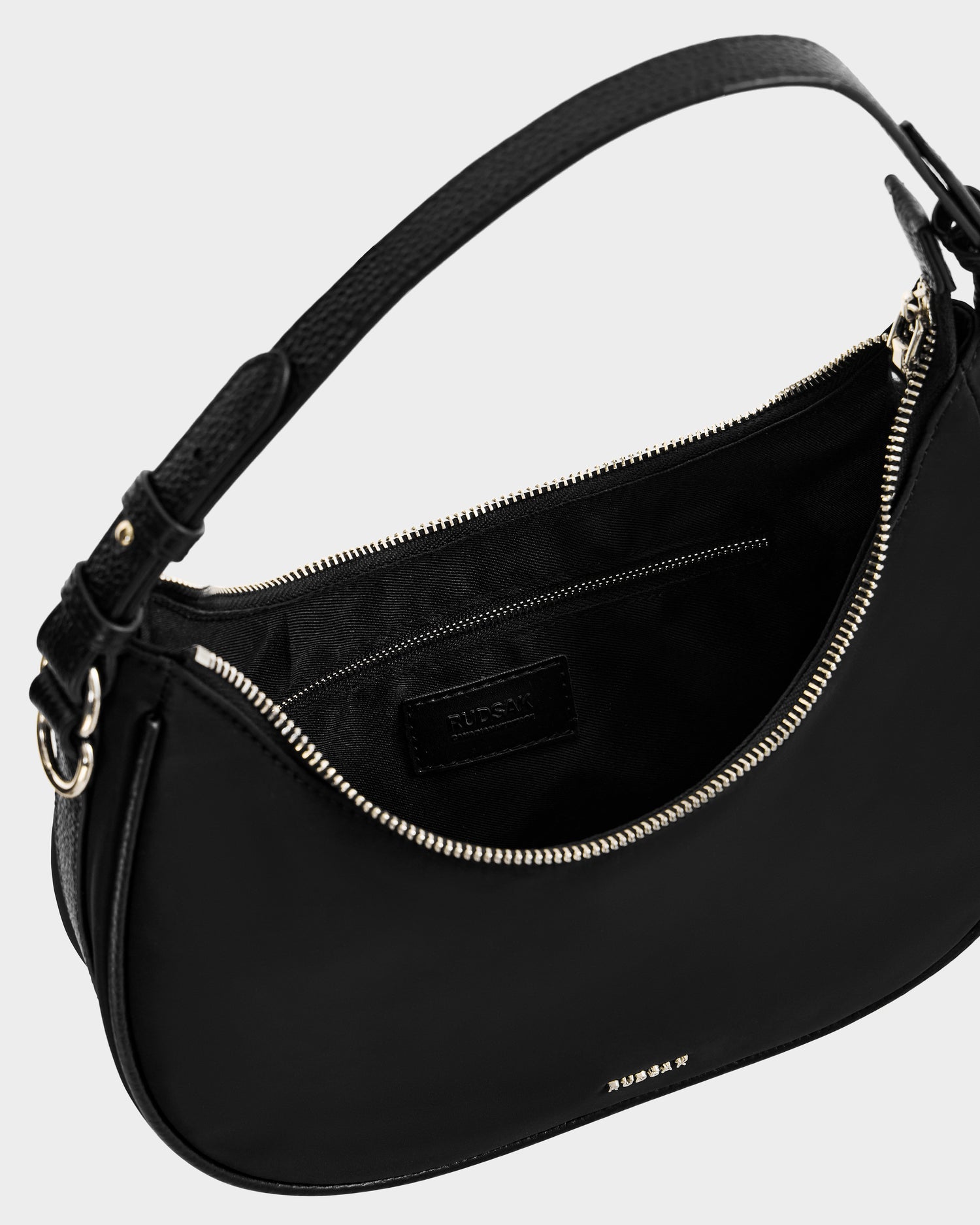 The NIKKI - L325512 Black by Rudsak is a black women’s crescent shoulder bag with a textured strap, gold-tone zipper, and interior pocket. The chic hobo silhouette features Rudsak’s name in gold on the exterior against a light background.