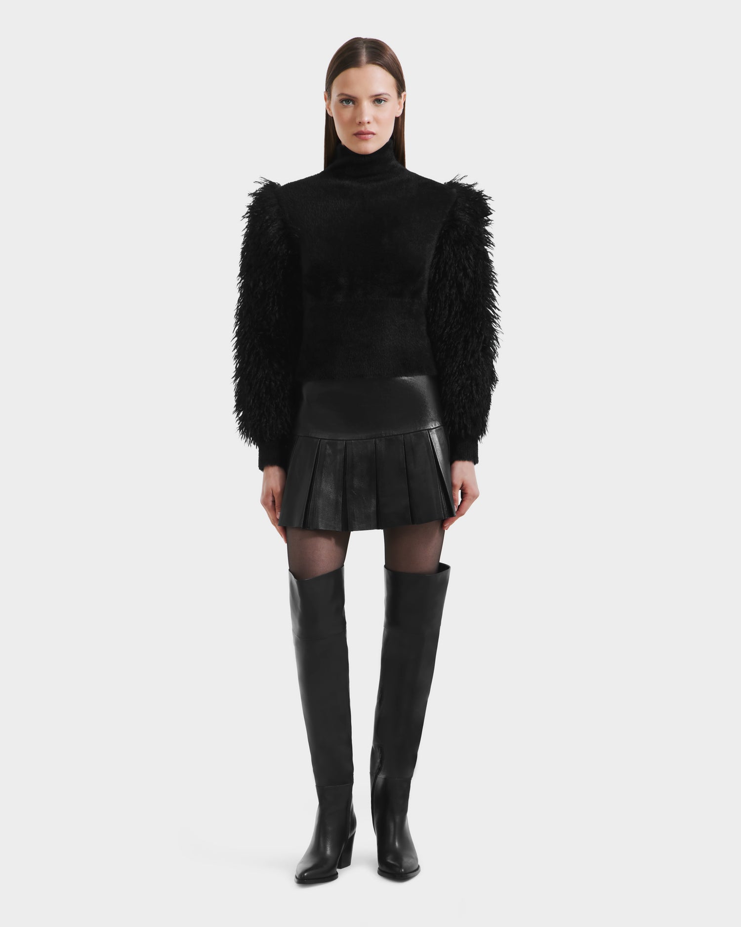 A woman stands facing forward in Rudsak’s NOEL - 8525571 Black mock-neck sweater with faux fur sleeves, styled with a black leather pleated mini skirt and over-the-knee boots—an elegant choice for winter layering against a white background.