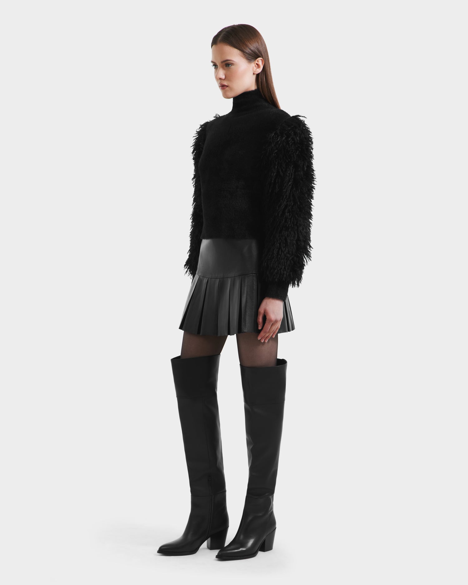 A woman stands against a plain background, wearing the Rudsak NOEL - 8525571 Black textured mock-neck sweater with faux fur sleeves, paired with a black pleated mini skirt, black tights, and knee-high heeled boots. She gazes slightly to the side.