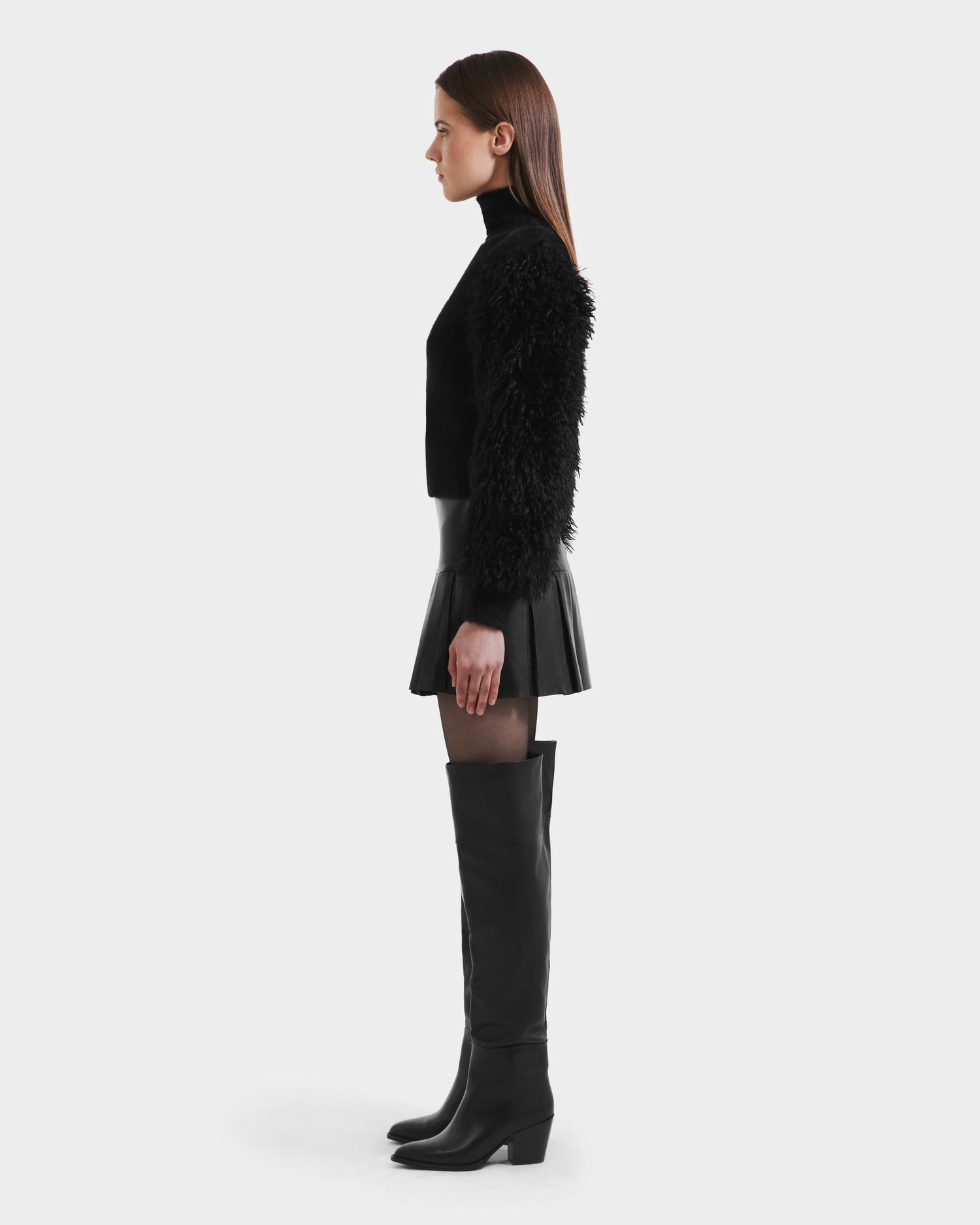 A woman stands in profile against a plain background, wearing the Rudsak NOEL - 8525571 Black mock-neck sweater with faux fur sleeves, paired with a black mini skirt, sheer tights, and knee-high boots for an elegant winter look.